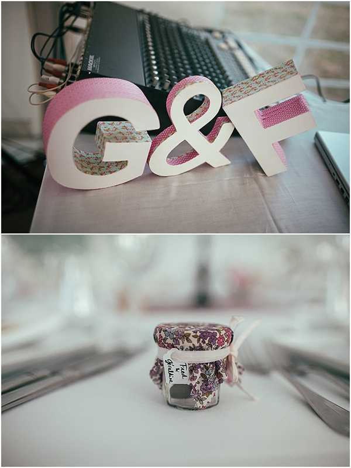 A two-image collage showing close-up detail shots of DIY-style wedding decor with a shabby-chic, crafty aesthetic. The top image shows freestanding white cardboard letters spelling 'G&F' decorated with pink polka-dot and floral Liberty-print washi tape, propped against a laptop on a table — likely preparation for the reception. The bottom image shows a small glass jar wedding favor placed on a set table, topped with a purple and mauve Liberty-style floral fabric circle secured with a white ribbon tied in a bow and a small handwritten label tag; place settings with silver cutlery and white plates are softly blurred in the background. The overall decor palette centers on pink, mauve, and floral Liberty-print fabrics, consistent with a vintage or crafted wedding styling theme. Both shots are close-up detail photographs with shallow depth of field.