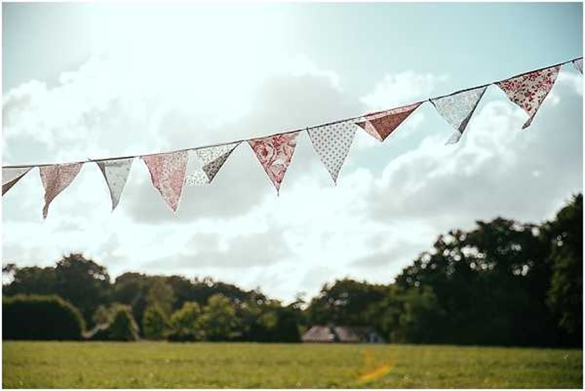 A detail shot of fabric bunting strung across an outdoor field setting, with the flags in sharp focus against a softly blurred rural background. The bunting features a mix of patterned fabrics including floral, polka dot, and plain triangular pennants in muted tones of dusty rose, sage green, and cream. The styling is consistent with a rustic or vintage-inspired wedding aesthetic. A farm building is faintly visible in the blurred background.