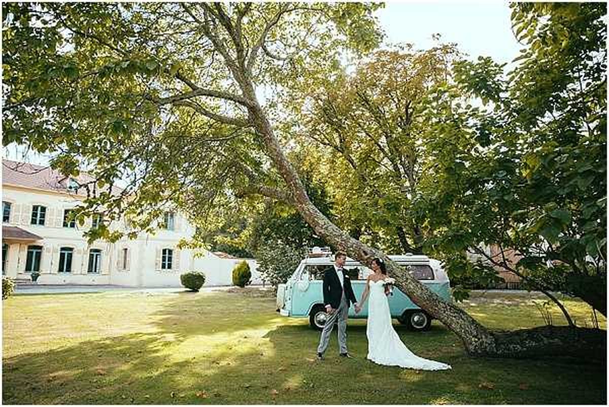 A bride and groom stand holding hands on the lawn of a French country estate, posing beside a vintage mint-green Volkswagen camper van. The bride wears a fitted white gown with a modest train, and the groom is dressed in a dark suit with light grey trousers. The setting is an outdoor garden in front of a classic white French manor house with shuttered windows. The composition is a wide shot that incorporates a large mature tree in the foreground, framing the couple and the decorative camper van as a styled prop. Potential venue feature image.