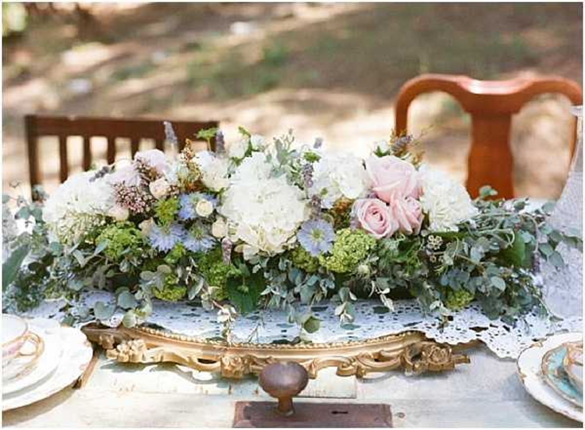 Close-up detail shot of an outdoor wedding reception table centerpiece arranged on an ornate gold baroque-style tray. The floral arrangement features ivory hydrangeas, blush pink roses, blue nigella, green viburnum, and trailing eucalyptus in a lush, garden-style composition. The table is dressed with a white lace doily runner, and mismatched vintage china place settings with floral detailing are visible at the edges of the frame. Wooden vintage chairs are visible in the soft-focus background, and the overall styling is romantic vintage with a soft blush, ivory, and sage green palette.