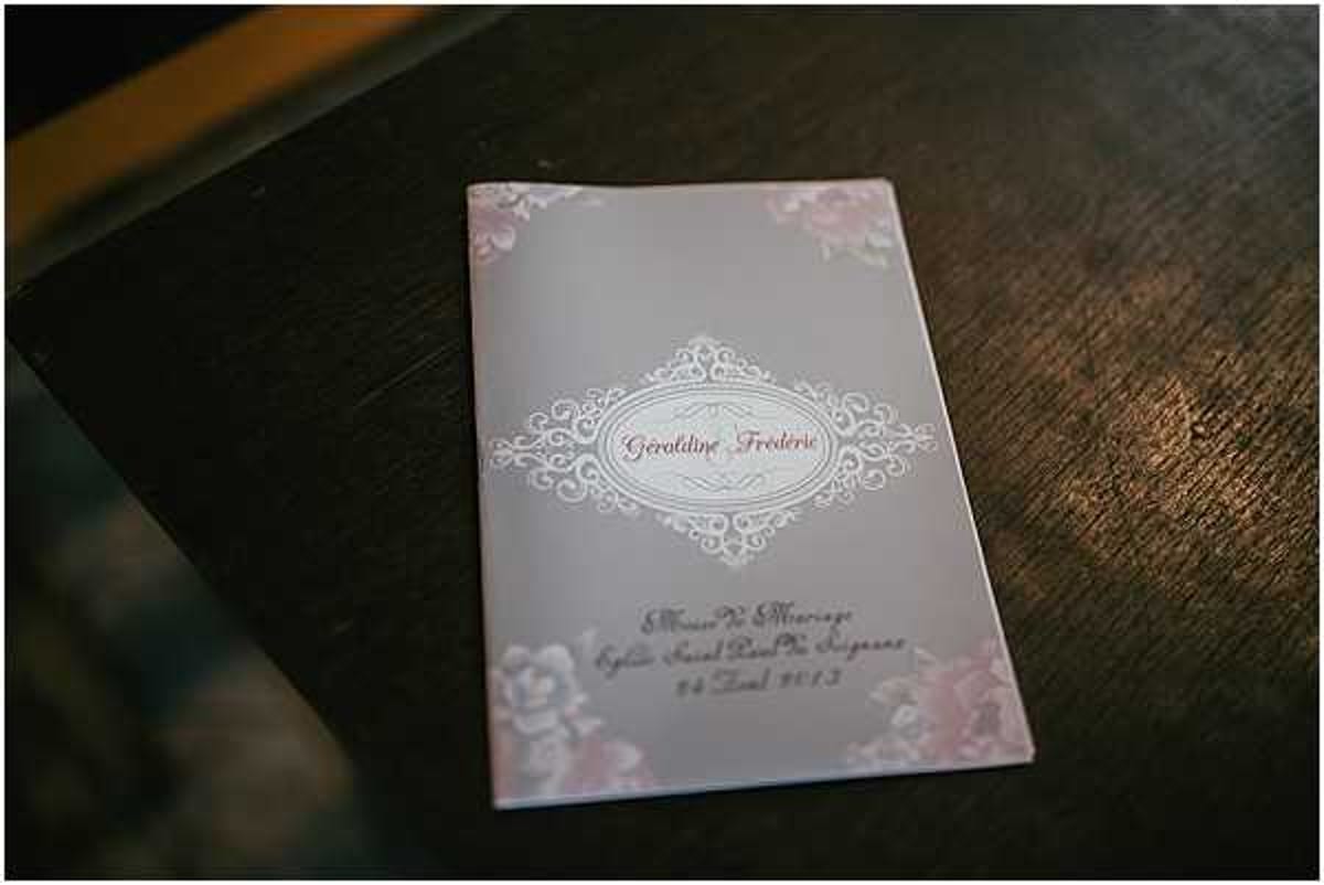 Close-up detail shot of a French wedding ceremony booklet resting on a dark wooden surface. The program is grey with soft blush pink floral corner embellishments and features an ornate white filigree frame enclosing the names 'Géroldine & Frédéric' in red script. Below the central motif, text reads 'Messe de Mariage, Eglise Saint Paul de Juignac, 14 Avril 2012,' indicating a Catholic church wedding ceremony. The design style is classic and romantic with a muted grey and blush pink colour palette.