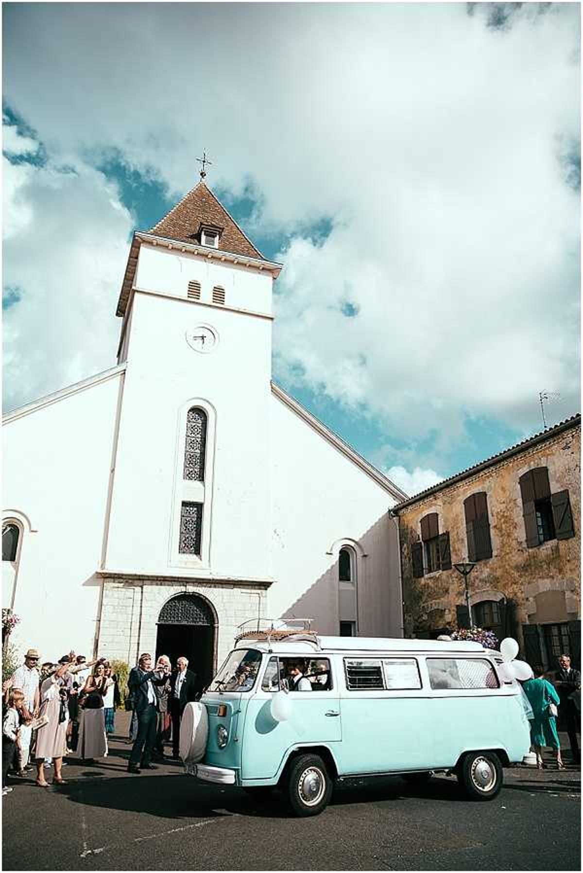 A wedding ceremony exit scene outside a white-rendered French village church with a square clock tower and arched doorway. A vintage mint-green Volkswagen Type 2 camper van, decorated with white balloons and flowers on the roof rack, is parked in the foreground as the couple departs inside it. Approximately 15–20 guests are gathered on the church steps and plaza, dressed in smart-casual attire including suits and summer dresses, some wearing hats. The styling has a retro-vintage feel, with the pale mint van serving as the wedding transport. Wide-angle shot taken from street level looking up toward the church tower.