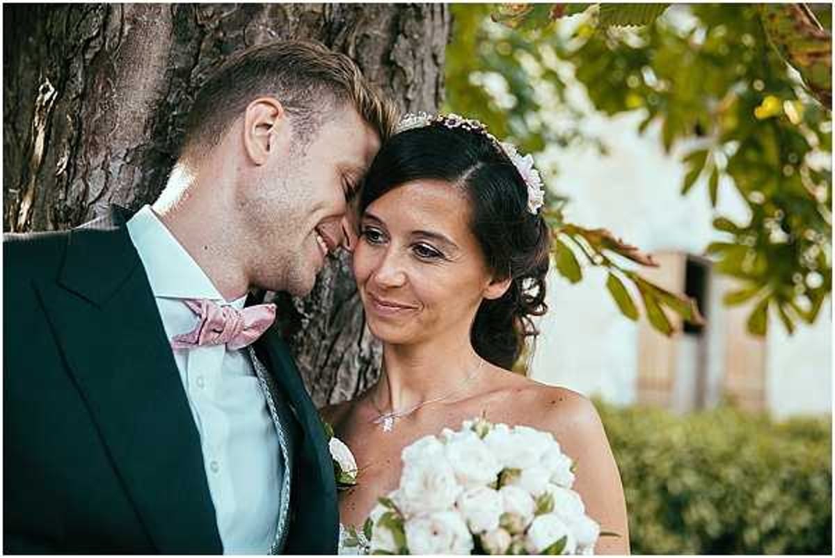 A close-up portrait of a bride and groom sharing an intimate moment outdoors, leaning their foreheads together beside a large tree trunk. The groom wears a dark navy suit with a light blue shirt and a dusty pink bow tie, while the bride wears a strapless white dress and a delicate floral-and-crystal hair accessory, with her dark hair styled in an updo. She holds a bouquet of white and blush garden roses and smiles warmly toward the camera. The styling palette is classic with soft pink and white accents.