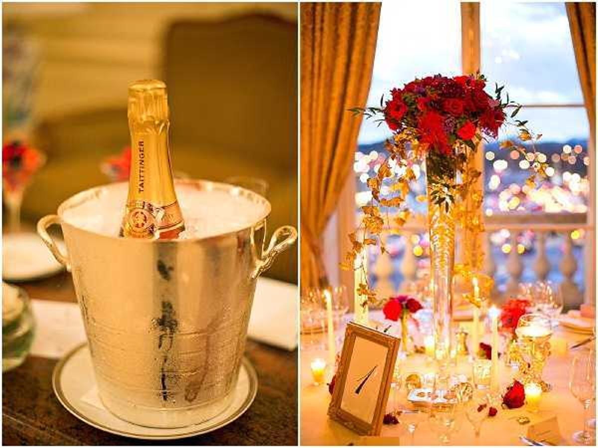 A split image showing two reception detail shots. On the left, a close-up of a Taittinger champagne bottle chilling in a polished silver ice bucket placed on a white plate atop a wooden table. On the right, a reception table styled in a red and gold palette featuring a tall glass trumpet vase centerpiece with red roses, gold trailing botanical accents, and warm fairy lights wound through the arrangement. The table is set with champagne flutes, pillar candles, scattered red rose petals, and a gold-framed table number card. Gold silk drape curtains frame a large window in the background, beyond which city lights are visible at dusk, suggesting an elevated indoor ballroom or hotel venue. The overall decor theme is classic and formal with a bold red and gold color scheme.