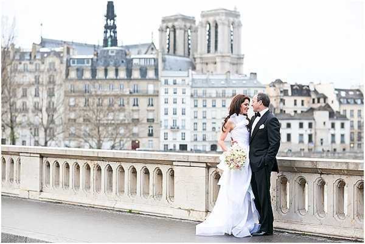 A wedding couple portrait taken outdoors on a Paris bridge, with the towers of Notre-Dame Cathedral and Haussmann-style buildings visible in the background. The bride wears a fitted white gown with a modest train and holds a bouquet of blush and ivory blooms, while the groom is dressed in a classic black tuxedo with a white dress shirt and bow tie. The two are close together in an intimate, almost-kissing pose, leaning against the ornate stone balustrade of the bridge. The image is a medium-wide portrait shot taken on an overcast day, with soft, diffused natural light.