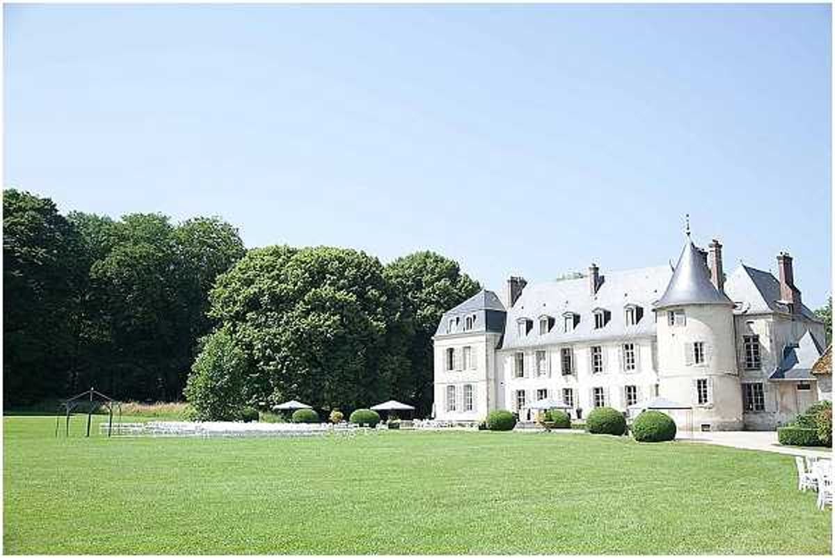 Wide exterior shot of a classic French chateau with white rendered facades, grey slate conical turret roofs, and symmetrical windows, set against an expansive manicured lawn. In the foreground, white ceremony chairs are arranged in rows on the grass alongside a simple wooden arbor structure, indicating an outdoor ceremony setup. Several white market umbrellas and what appears to be cocktail furniture are positioned near the chateau entrance. No people are visible in the image. Potential venue feature image.
