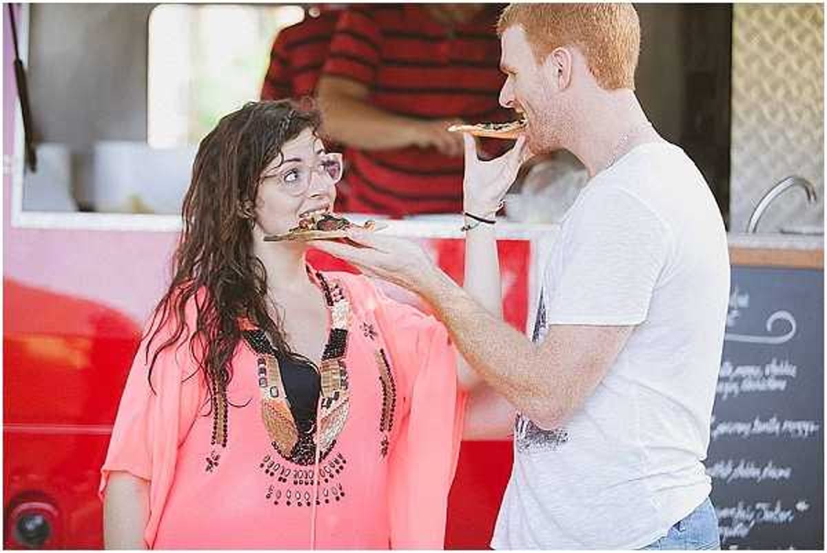 A candid outdoor shot of a man and woman eating pizza or flatbread slices near a red food truck or catering vehicle, likely during a wedding cocktail hour or reception. The woman wears a coral/salmon pink kaftan-style cover-up with black and gold beaded embroidery detail, while the man wears a white t-shirt and jeans. A chalkboard menu sign is partially visible on the right side of the frame, and a third person in a red striped shirt is visible in the background behind the food truck counter. The image is a medium close-up portrait-style shot with a relaxed, casual atmosphere suggesting a boho or informal wedding reception style.