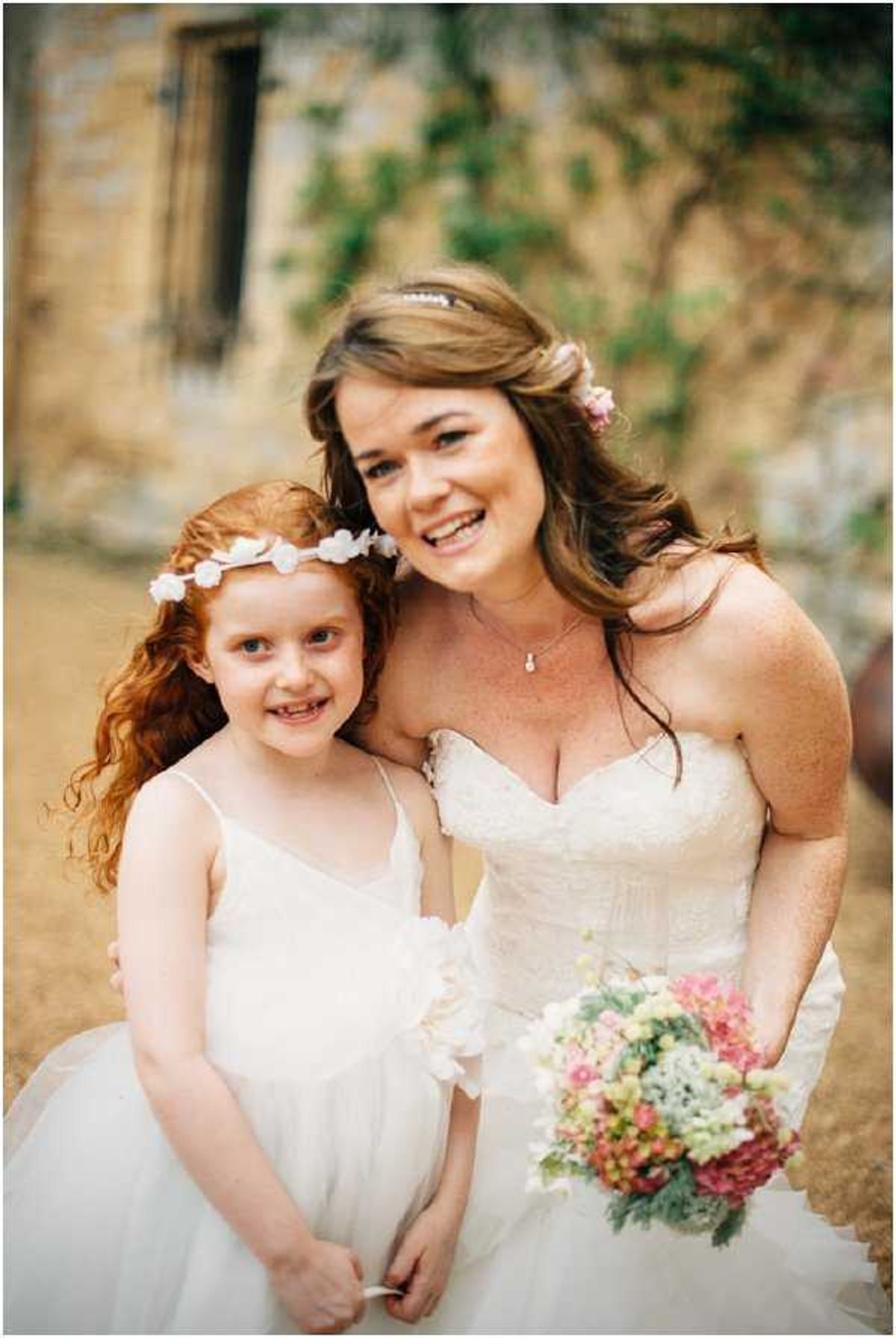 A portrait of a bride posing with a young flower girl outdoors in front of a stone building with ivy. The bride wears a strapless lace corset-style wedding gown and holds a loose bouquet of pink hydrangeas, pale green foliage, and small pastel blooms; she has small pink flowers tucked into her half-up brown hair. The flower girl wears a simple white spaghetti-strap dress and a white floral crown in her curly red hair. The styling has a relaxed, rustic feel. Close-up portrait shot with shallow depth of field.