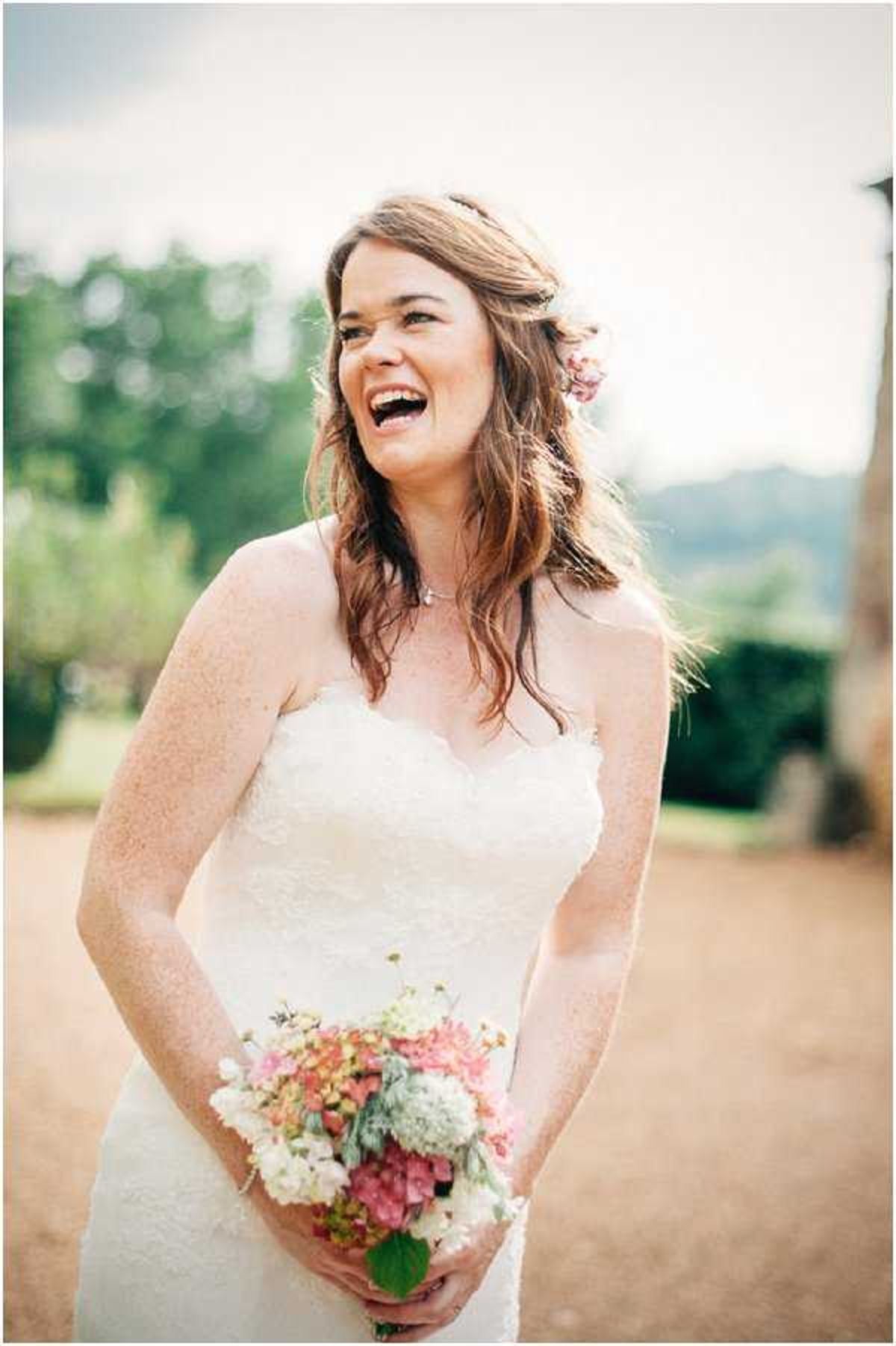 A bridal portrait taken outdoors, showing the bride laughing and looking to one side. She wears a strapless white lace dress with a sweetheart neckline and holds a loosely arranged bouquet of white hydrangeas, pink hydrangeas, and small filler flowers with green foliage. Her auburn hair is worn down in soft waves with small pink flowers tucked behind her ear, and she wears a delicate necklace. The background is blurred, suggesting a garden or grounds setting with trees and open landscape. The shot is a close-up portrait with a shallow depth of field.