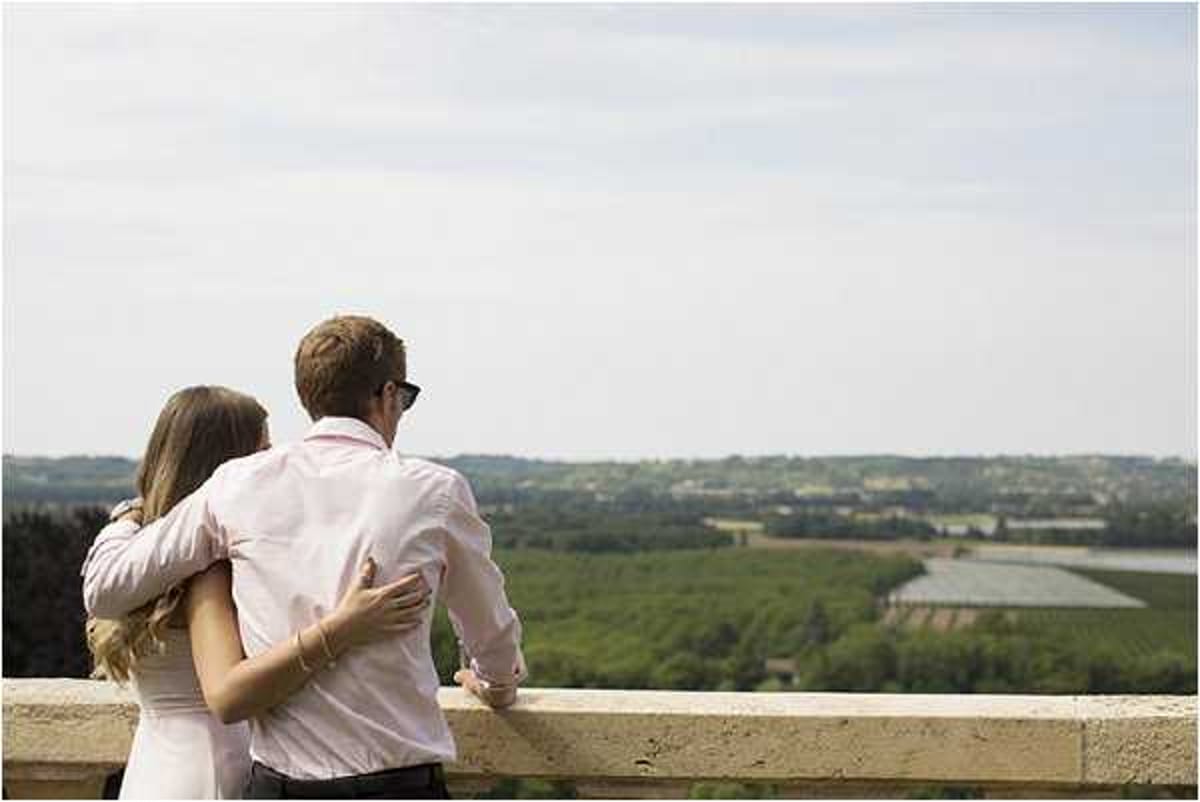 A couple stands together on a stone balustrade terrace, viewed from behind, looking out over an expansive landscape of vineyards and fields. The woman wears a white dress and the man wears a light pink dress shirt with dark trousers and sunglasses, his arm around her shoulders. The wide shot captures the couple in an intimate, relaxed pose against the panoramic countryside backdrop. The setting suggests a chateau or elevated venue with a formal stone balcony railing.