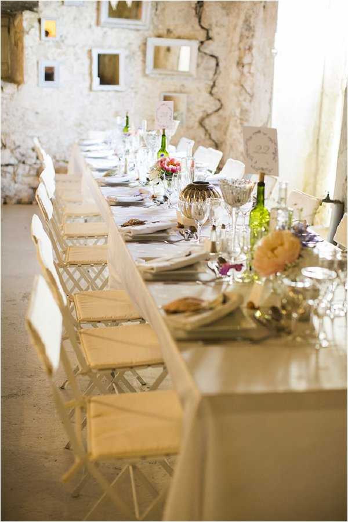 A reception tablescape detail shot showing a long banquet table set for a wedding dinner inside a rustic stone-walled venue with exposed, cracked plaster walls. The table is covered in a white linen cloth and lined with white painted wooden folding chairs with woven seats on both sides. Centerpieces include coral and pink ranunculus or peonies, small purple florals, green wine bottles used as vases, mercury glass and metallic vessels, crystal glassware, and white table number cards. Empty white ornate picture frames of varying sizes are mounted decoratively on the distressed stone wall in the background, reinforcing a shabby-chic or vintage-rustic styling theme. The perspective is a low-angle shot running the length of the table, giving a sense of scale to the long setup.