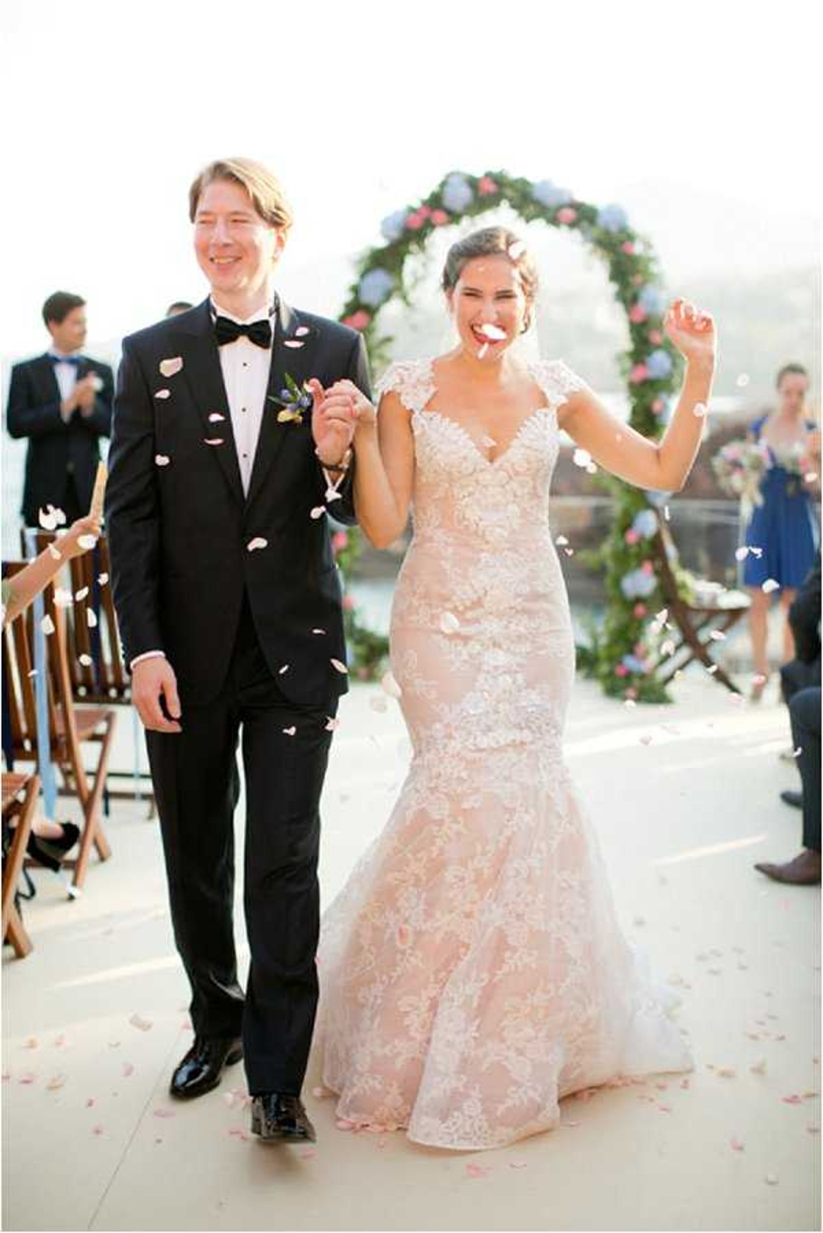 The bride and groom walk back down the aisle as a newly married couple during an outdoor ceremony recessional, with guests tossing pink flower petals over them. The groom wears a black tuxedo with a black bow tie and a small floral boutonniere, while the bride wears a blush pink lace mermaid-style gown with a V-neckline and cap sleeves. Behind them is a large circular floral arch decorated with pink roses, white blooms, and greenery, set against an open outdoor terrace with what appears to be a water view in the background. Guests, including women in cobalt blue dresses, are visible on either side of the aisle lined with wooden folding chairs; this is a portrait-style, mid-length shot capturing the couple's joyful expressions.