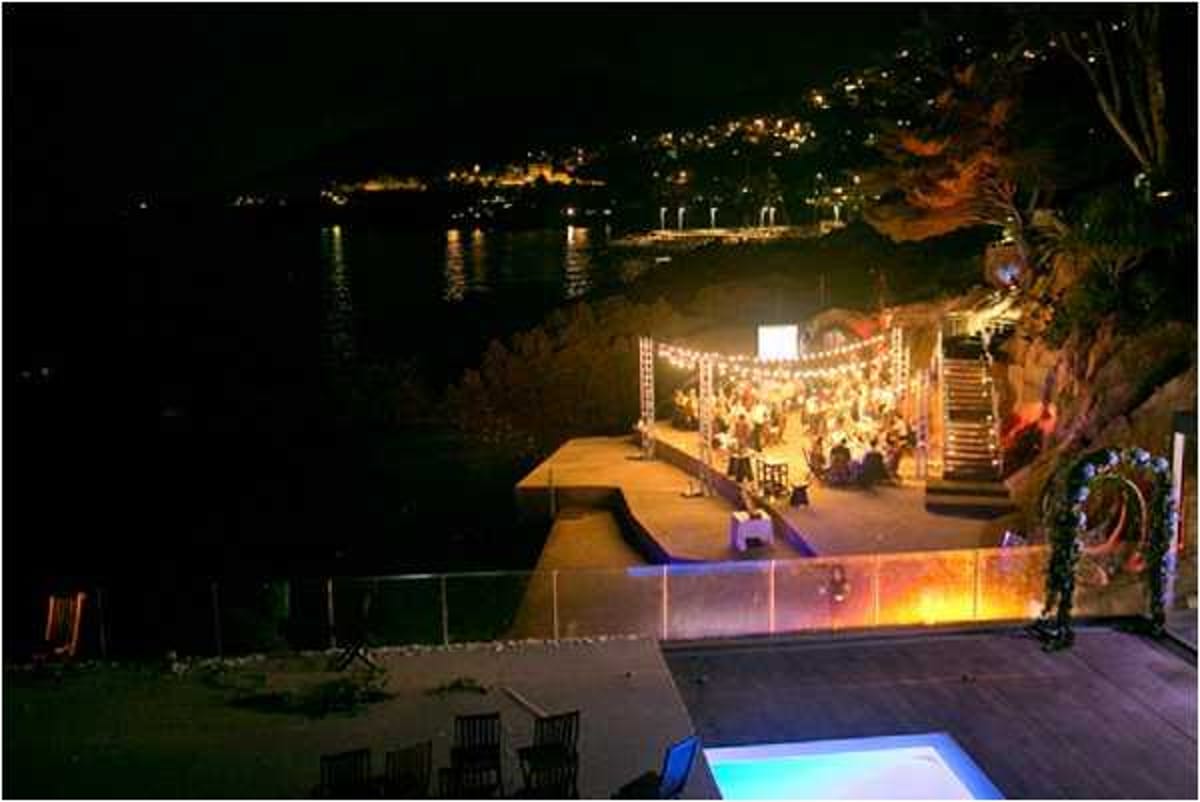 An aerial wide shot of an outdoor wedding reception taking place at night on a waterfront terrace or pier, with a lit harbor and town visible in the background across the water. The reception area is strung with warm festoon/bistro lights suspended on a truss structure, illuminating a large gathering of guests seated at round tables. A projection screen is visible at one end of the space, and the overall decor palette appears warm and golden from the lighting. In the foreground, a swimming pool with blue lighting and garden seating area are visible on a lower terrace level. The setting appears to be a private coastal villa or waterfront venue, with the event area positioned directly beside the water's edge. Potential venue feature image.