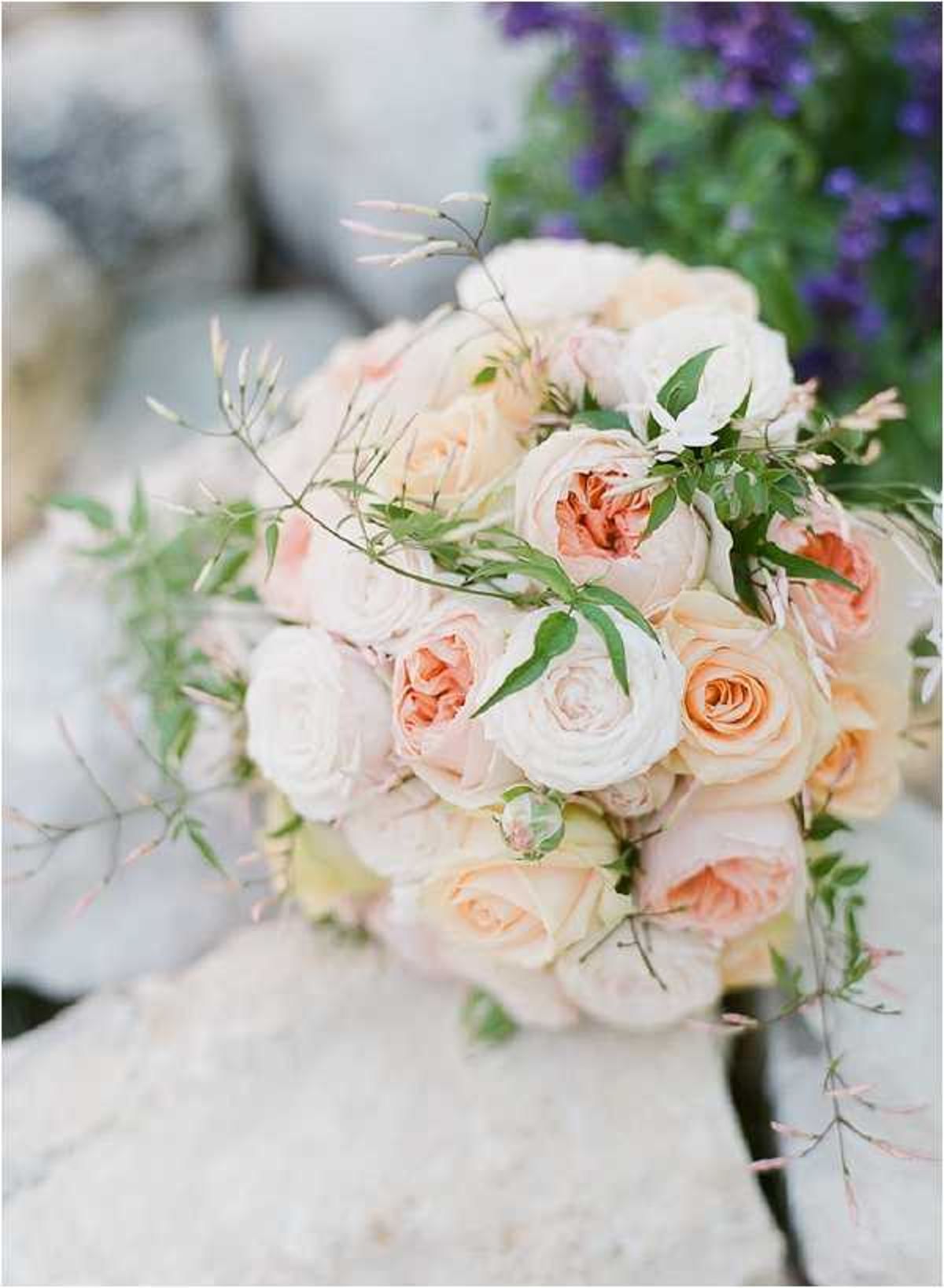 Close-up detail shot of a bridal bouquet resting on a pale stone surface outdoors. The bouquet is composed of garden roses and peonies in blush pink, peach, and cream tones, accented with small green leaves, delicate wispy stems, and a single closed bud. Trailing greenery extends loosely from the arrangement, giving it an organic, unstructured shape. Purple flowering shrubs are softly blurred in the background, adding a contrasting color note to the composition.