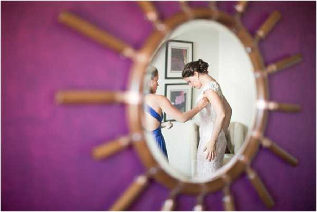 A getting-ready scene captured as a reflection in a sunburst-style oval mirror with a wooden spoke frame, mounted on a vibrant magenta-purple wall. The reflection shows a bride in a fitted lace gown with cap sleeves having her dress fastened by a helper dressed in a cobalt blue strapless dress. Two framed artworks are visible on the white wall behind the pair in the reflection. The composition uses the mirror as a creative framing device, with the mirror itself in sharp focus while the surrounding room is intentionally blurred, creating a portrait-style detail shot.