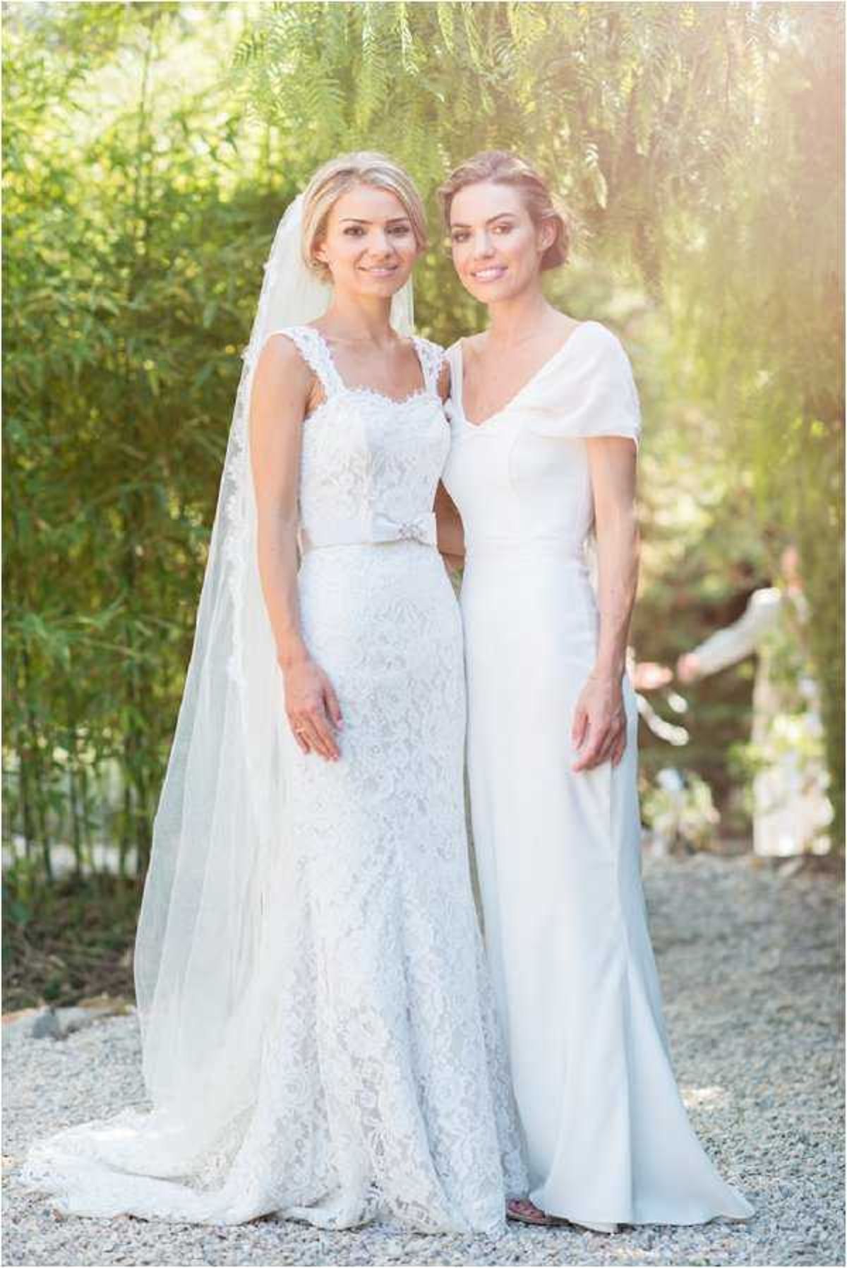 A portrait of two women posing together outdoors on a gravel path, shot in warm natural light with a softly blurred garden background. The bride on the left wears a fitted white lace gown with cap-style lace straps, a small bow detail at the waist, a cathedral-length veil, and a long train; her blonde hair is styled in a loose updo. The woman on the right, likely a bridesmaid or close family member, wears a sleek ivory short-sleeved gown with a V-neckline and minimal detailing, with her dark hair in an updo. The overall styling is classic and polished, with a predominantly white and ivory palette.