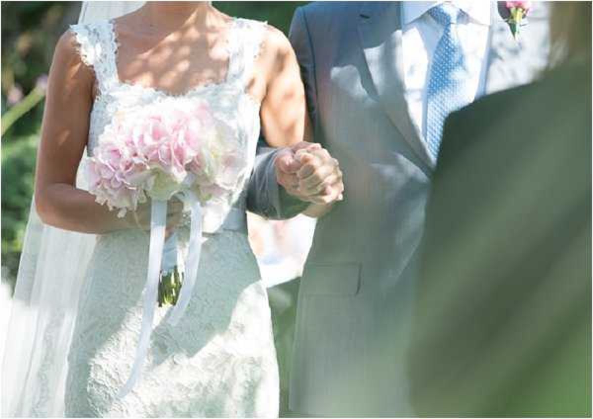 A close-up portrait shot of a bride and groom holding hands during what appears to be an outdoor ceremony. The bride wears a white lace dress with delicate ruffle or floral strap detailing and holds a bouquet of soft pink and cream hydrangeas tied with long white satin ribbons. The groom wears a light grey suit with a light blue polka-dot tie and has a small pink floral boutonniere on his lapel. The image is brightly lit with natural sunlight, giving it a soft, high-key look.