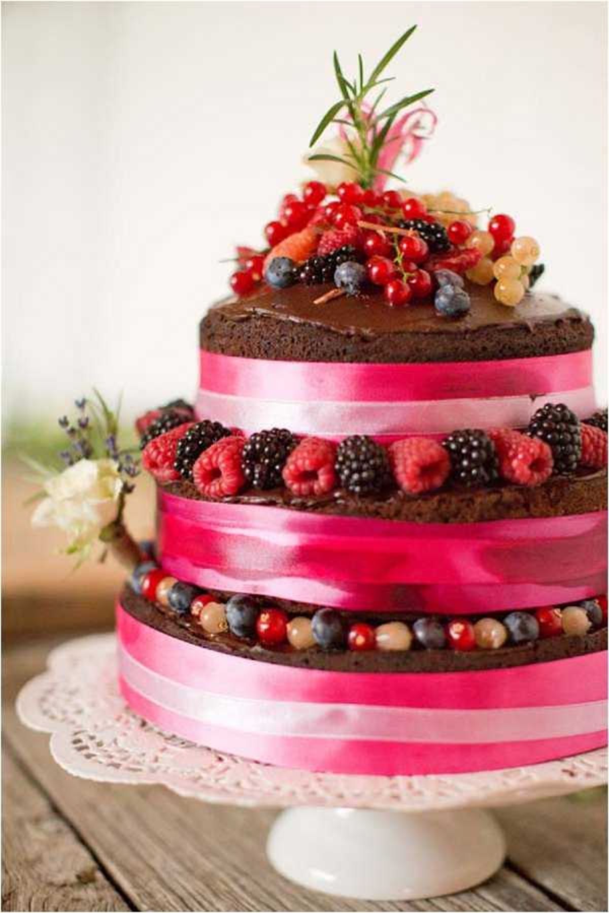 Close-up detail shot of a three-tier naked chocolate wedding cake displayed on a white ceramic cake stand with a white doily, set on a rustic wooden surface. Each tier is wrapped with wide hot pink satin ribbon and decorated between layers with fresh berries including raspberries, blackberries, blueberries, red currants, and white currants. The top tier is finished with a generous cluster of mixed berries, red currants, white currants, and a sprig of fresh rosemary. A small arrangement of cream-colored flowers and lavender is tucked beside the base tier. The overall styling is rustic with a bold berry and hot pink color palette.