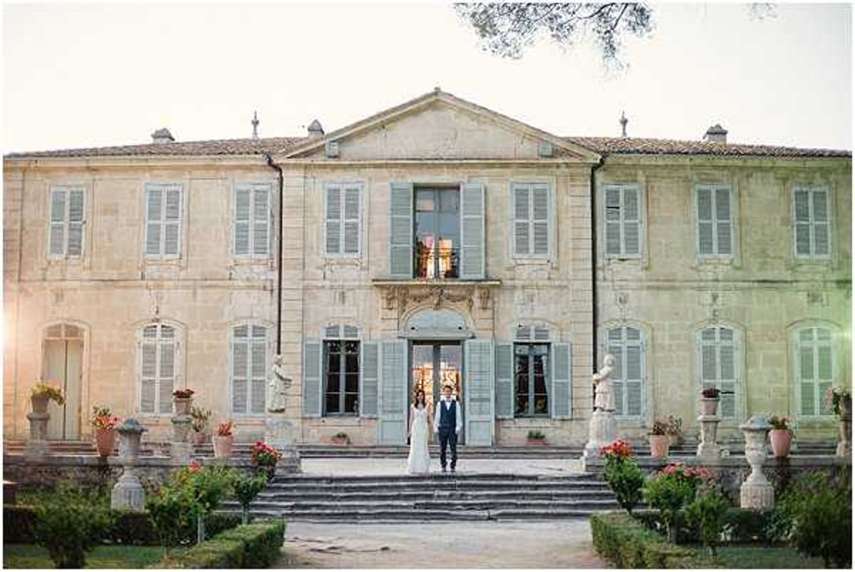 A couple poses together on the front steps of a large French chateau, standing centered in front of the main entrance doorway. The bride wears a long white gown and the groom is dressed in a navy suit with no tie. The chateau is a two-story stone building with pale blue-grey shutters, stone statues flanking the steps, terracotta planters with red flowering plants, and a symmetrical formal facade. The shot is taken at dusk with warm golden light visible at the horizon. Wide portrait shot capturing the full building exterior and formal garden foreground. Potential venue feature image.