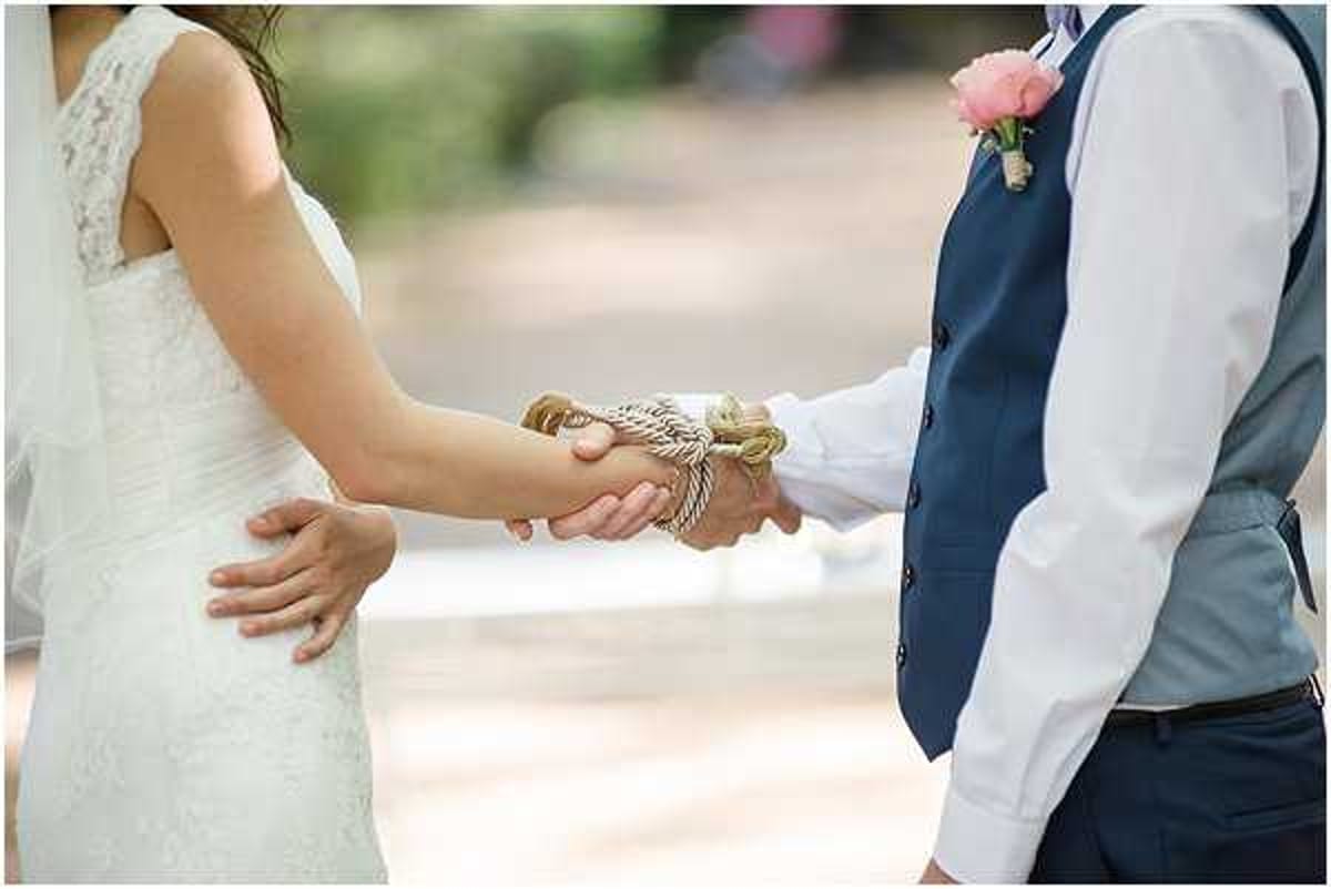 Close-up detail shot of a handfasting ceremony, showing the joined hands of a bride and groom bound together with a twisted cream and gold rope or cord. The bride is wearing a white lace dress with a sheer veil visible at her side, and a third hand rests on her back. The groom is dressed in a navy blue vest over a light blue dress shirt with navy trousers, and wears a coral/pink boutonniere on his lapel. The outdoor ceremony setting is softly blurred in the background. The composition focuses tightly on the hands and the binding cord, emphasizing this ritual element.