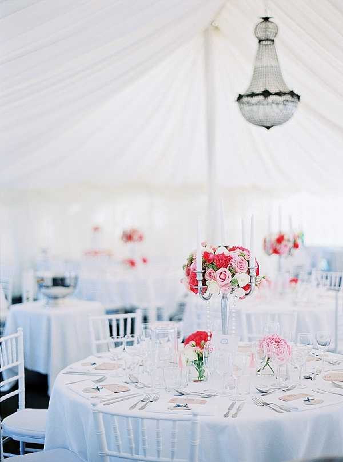 A wedding reception setup inside a white draped marquee tent, shot from a medium-wide angle showing multiple round tables dressed in white floor-length linens with white chiavari chairs. The foreground table features a tall silver candelabra centerpiece topped with a compact arrangement of hot pink and blush pink roses, with smaller bud vases holding pink peonies and red roses scattered across the table. Place settings include silverware, multiple wine glasses, and kraft paper place cards with small blue ribbon accents. Additional matching tall candelabra centerpieces with hot pink and blush floral arrangements are visible on tables in the background. A crystal chandelier hangs from the tent ceiling, which is lined with white draped fabric. The overall decor palette is white, hot pink, and blush pink in a classic style.