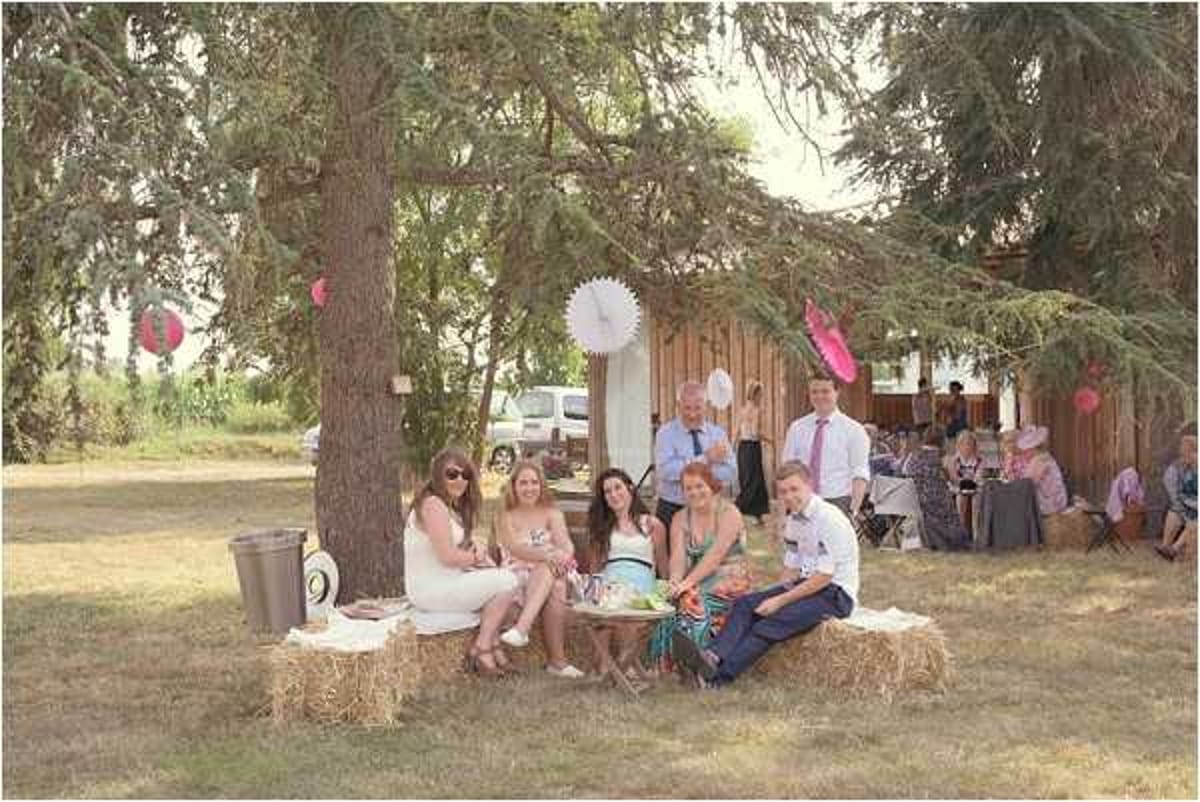 An outdoor wedding cocktail hour or reception gathering on a lawn, with a group of approximately six guests seated casually on hay bales around a small low table, with additional guests visible in the background seated in chairs near a rustic wooden structure. The decor includes white and pink paper fan pompoms hung from tree branches, contributing to a relaxed, rustic-boho styling theme. Guests are dressed in summer attire including a white dress, floral prints, and light-colored suits with bow ties. The overall composition is a medium-wide candid shot capturing the informal, relaxed atmosphere of the outdoor celebration.