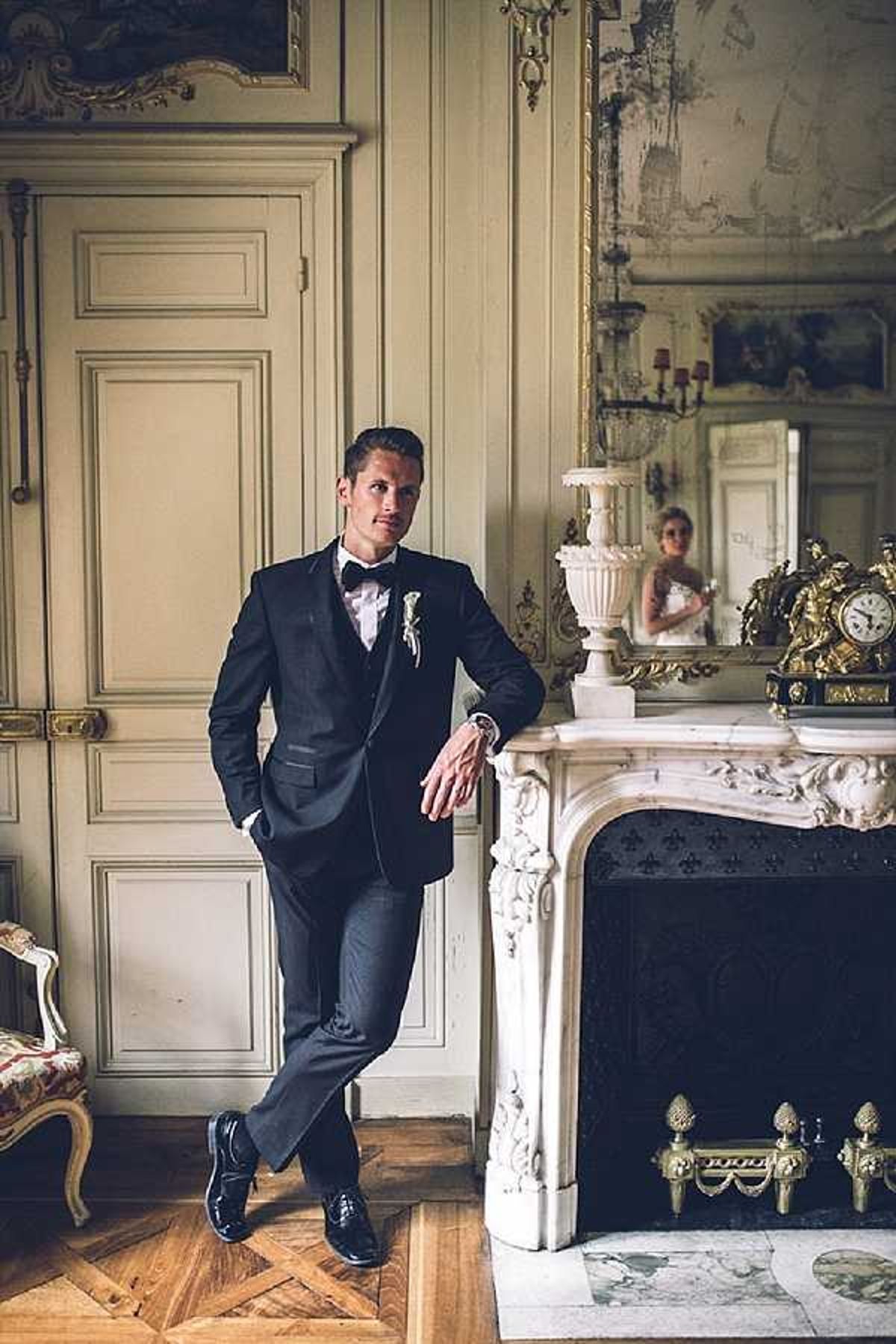 A groom stands leaning against an ornate white marble fireplace inside what appears to be a French chateau, posing for a portrait in an elaborately decorated room with cream-paneled walls, herringbone parquet floors, and gilt-framed artwork. He is dressed in a dark navy or black tuxedo with a black bow tie, white dress shirt, and a white floral boutonniere, completed with black patent leather shoes and a watch. The mantelpiece is decorated with a large white marble urn and an ornate gold clock, with a large gilt-framed mirror above reflecting the bride in a white dress in the background. The composition is a full-length portrait with warm, slightly moody lighting that highlights the classic French interior decor alongside a Louis XVI-style armchair visible at the left edge of the frame.