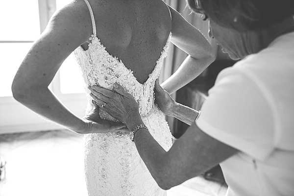 A black-and-white close-up portrait of a bride being helped into her wedding dress by another person, likely a mother or family member. The image is shot indoors with soft natural light coming from a window in the background. The bride's dress features thin spaghetti straps and intricate lace appliqué detailing across the back, with a deep V-back silhouette. The assistant's hands, adorned with a bracelet and ring, are visible fastening or adjusting the back of the gown. The composition is a tight detail shot focused on the backs and hands, emphasizing the texture of the lace against high-contrast monochrome tones.