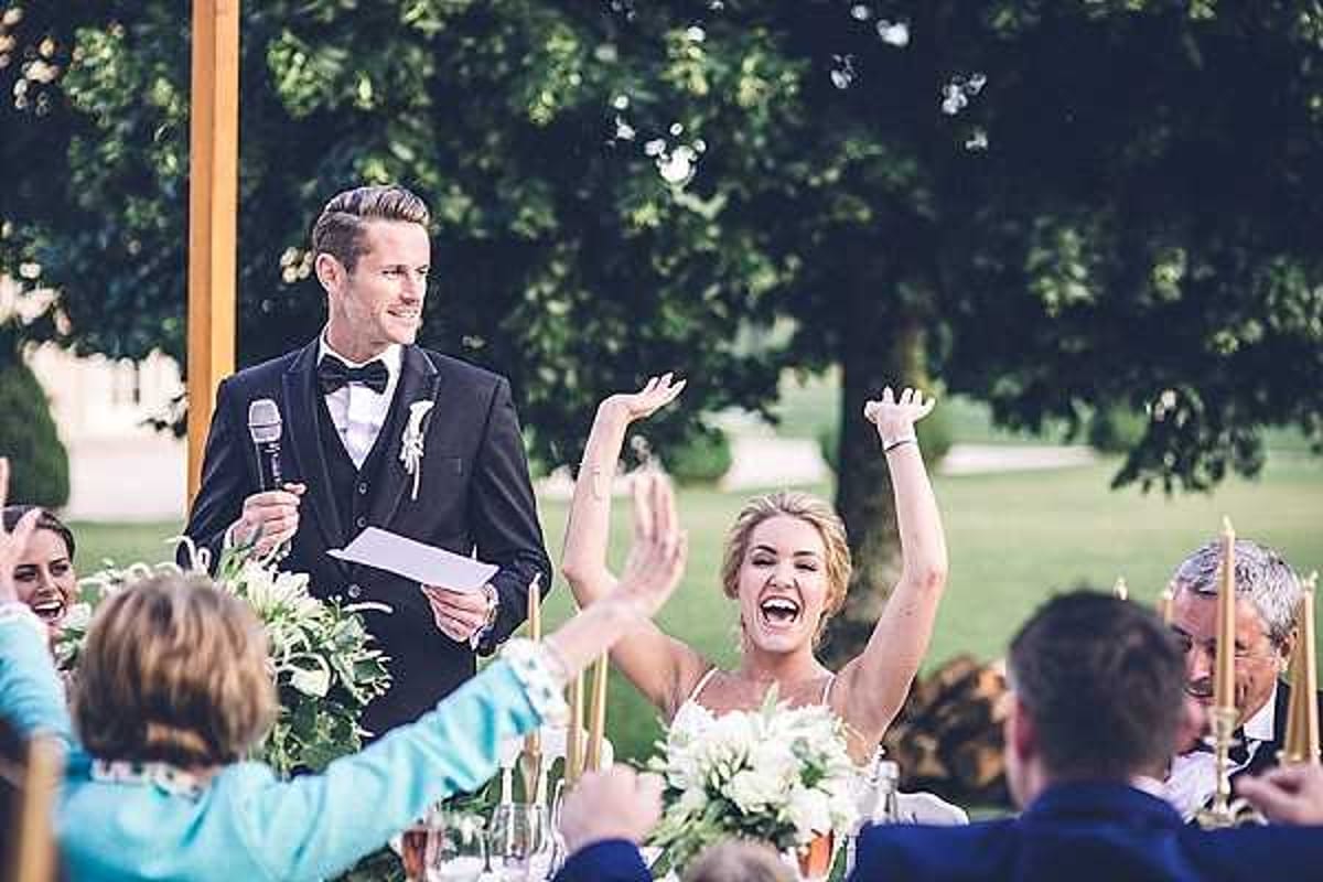 A speech is taking place during an outdoor wedding reception, with the groom standing and holding a microphone and papers while wearing a navy tuxedo with a black bow tie and a white boutonniere. The bride, seated beside him in a white spaghetti-strap dress with an updo hairstyle, is laughing with both arms raised in an animated, joyful reaction. Guests seated around the long reception table are also laughing and raising their hands, visible in the foreground and background. The table is decorated with white and green floral centerpieces and tall gold taper candles, set against a lush green garden backdrop in what appears to be a classic French estate setting. Wide candid portrait shot capturing a lively, celebratory moment.