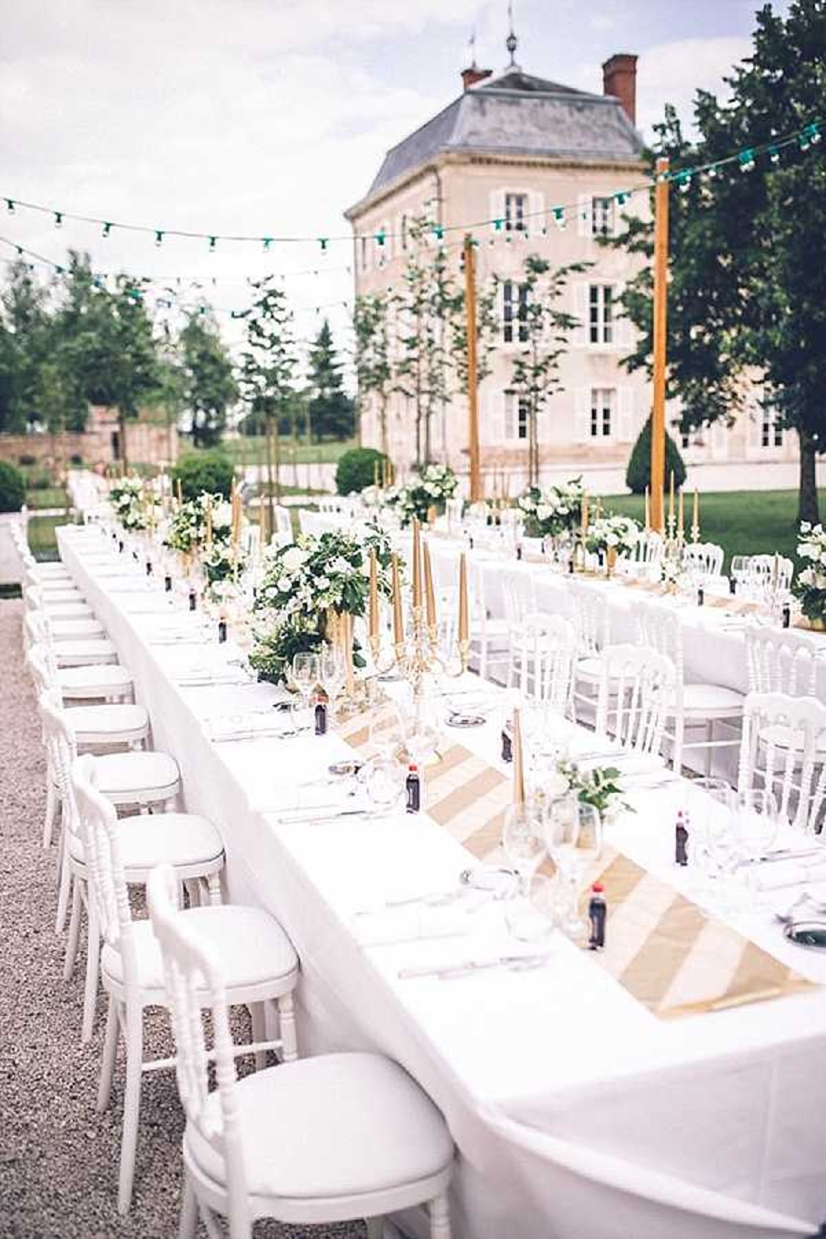 An outdoor wedding reception table setup photographed in a wide shot from one end of a long banquet-style table, with a French chateau visible in the background. The table is dressed in white linen with gold and white diagonal-striped table runners, set with glassware, silverware, and individual place settings. Centerpieces consist of tall gold candelabras holding slender gold taper candles, interspersed with low floral arrangements of white blooms and greenery. White Chiavari-style chairs line both sides of the table. Teal pennant bunting and string lights are strung overhead between tall gold poles, adding a festive element to the classic white and gold decor palette. No guests are present in the image. Potential venue feature image.