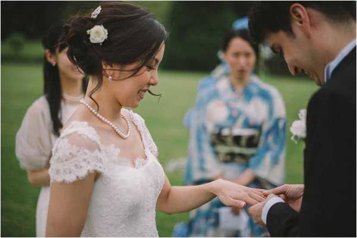 The ring exchange moment during an outdoor wedding ceremony, with the groom placing a ring on the bride's finger as she smiles. The bride wears a lace cap-sleeve wedding dress with a sweetheart neckline, a pearl necklace, and has a white rose hair accessory in her dark updo. In the background, two guests are visible — one in a beige dress and another wearing a blue-and-white floral kimono, suggesting a blend of French and Japanese cultural elements. The shot is a medium close-up portrait focused on the couple's hands and the bride's expression, with the background softly blurred.