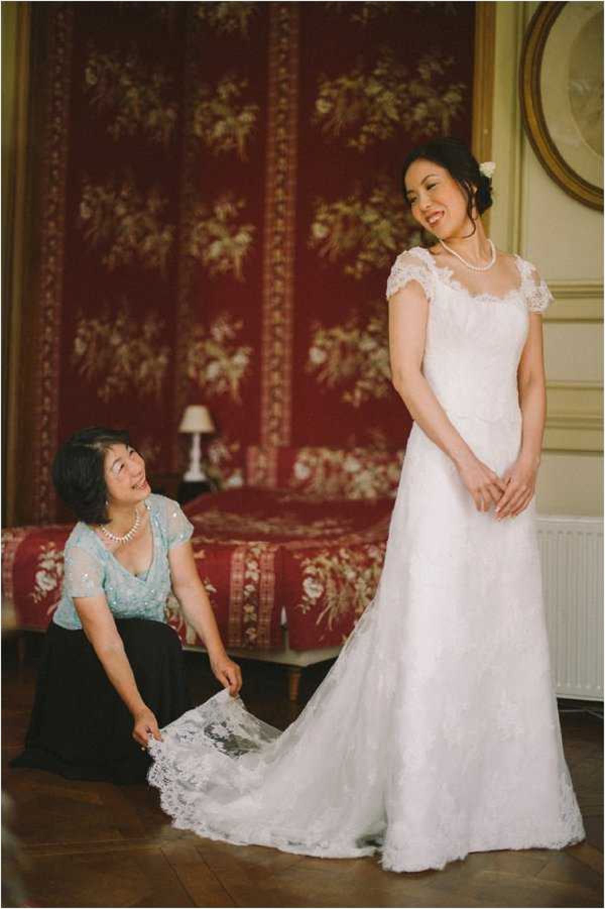 A getting-ready moment captured indoors in what appears to be a chateau or manor house bedroom, featuring ornate red floral wall panels and gold-framed artwork in the background. The bride stands in a white lace cap-sleeve gown with a lace train, wearing a pearl necklace and a white floral hair accessory, smiling down at a woman kneeling beside her who is adjusting and arranging the lace train. The second woman, likely the bride's mother, wears a light blue sequined top and black skirt with a pearl necklace. The composition is a medium portrait shot with warm, slightly vintage toning, taken on dark hardwood floors.