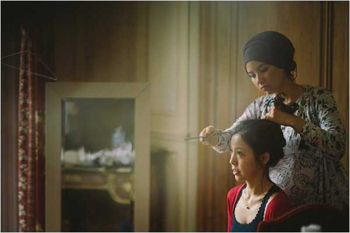 A getting-ready scene showing a hair stylist wearing a dark head wrap and a floral-print top working on a seated woman's updo hairstyle, using what appears to be bobby pins or hair pins. The seated woman is dressed in a red and navy top. The setting appears to be an indoor room with warm, dim ambient lighting, sheer curtains diffusing natural light from a window, and an ornate mirror reflecting a decorative fireplace mantel in the background, suggesting a chateau or historic venue interior. The image is a medium portrait shot with a shallow depth of field and warm, slightly dark tones.