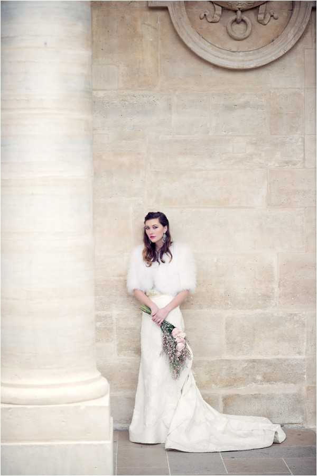 A bridal portrait taken outdoors against a pale limestone ashlar wall with classical architectural details, including a large stone column in the foreground and a carved stone medallion relief above. The bride stands alone, wearing a fitted ivory gown with a train and a cream satin waist sash, layered with a white fluffy faux-fur or marabou feather cropped bolero shrug. She holds a loosely cascading bouquet of blush pink roses and trailing baby's breath at her side. Her dark hair is styled up, and she wears statement drop earrings with bold red lipstick. The composition is a full-length portrait shot with a soft, slightly hazy quality and muted, warm tones consistent with a vintage-inspired or art deco styling theme.