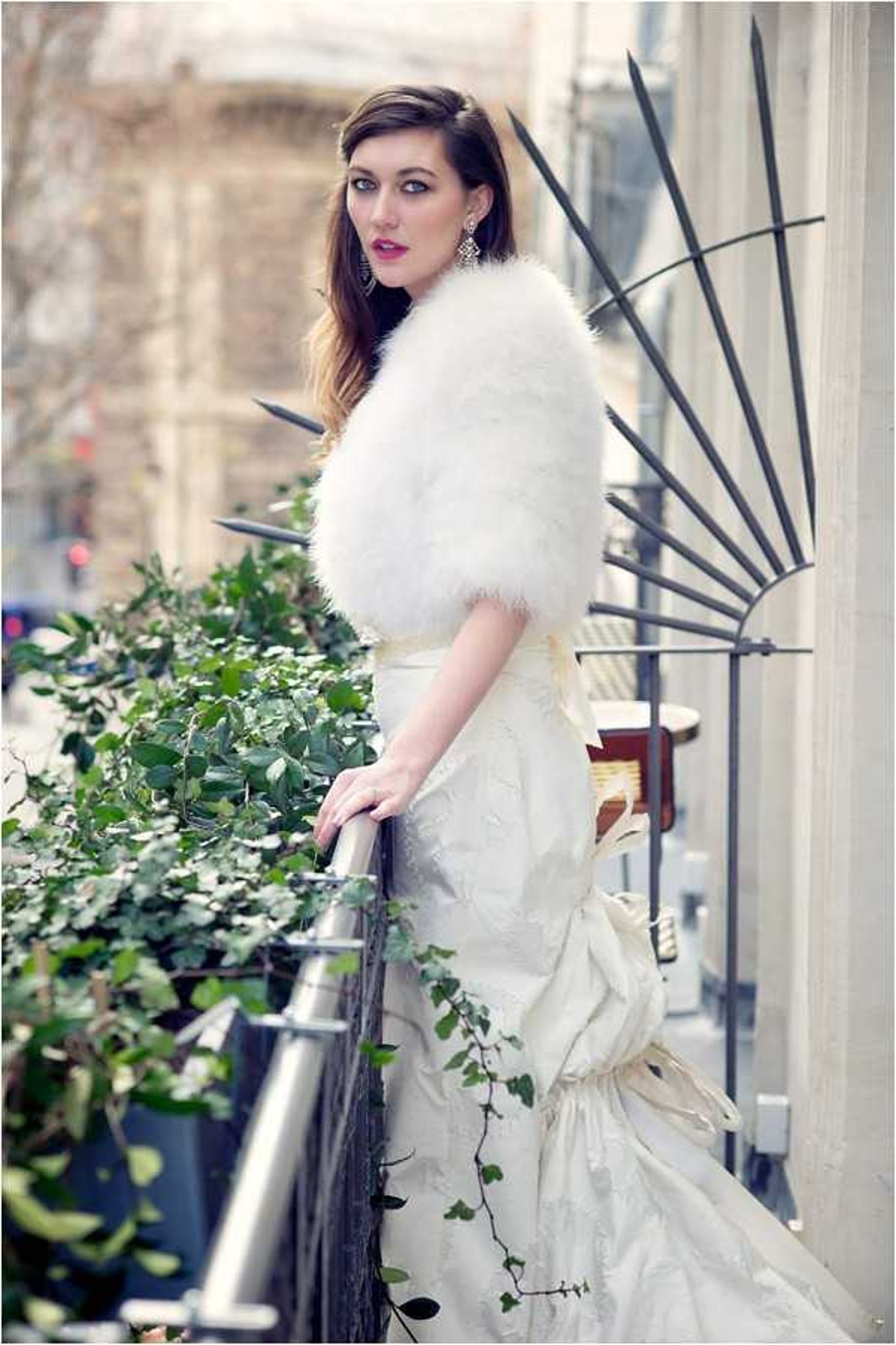 A bridal portrait taken on an outdoor urban balcony or terrace, with a city street visible in the blurred background. The bride stands in a three-quarter pose, wearing an ivory textured gown with a large bow detail at the back and a white feather or faux-fur stole draped over her shoulders. She accessorizes with chandelier earrings and wears bold red lipstick, with her dark hair swept to one side. The balcony railing features green ivy and boxwood planters, and a decorative black metal fan-shaped architectural element is visible on the building facade. The overall styling has a classic Parisian aesthetic. Portrait composition with a shallow depth of field.