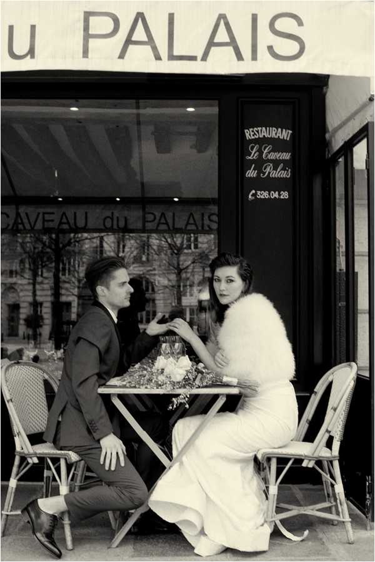 A black-and-white portrait of a bride and groom seated at a small outdoor café table in front of Restaurant Le Caveau du Palais, a Parisian establishment visible by its awning and signage. The groom, wearing a dark suit with a tie, leans forward toward the bride, whose hand he holds across the table; he is seated in a classic French bistro rattan chair with one leg extended. The bride wears a fitted white gown and a fluffy fur stole over her shoulders, with her dark hair styled in an updo, and she glances back toward the camera. A small floral arrangement and what appear to be champagne flutes sit on the bistro table, and the composition is a medium full-length portrait with high contrast tones giving the image a vintage, mid-century Parisian feel.