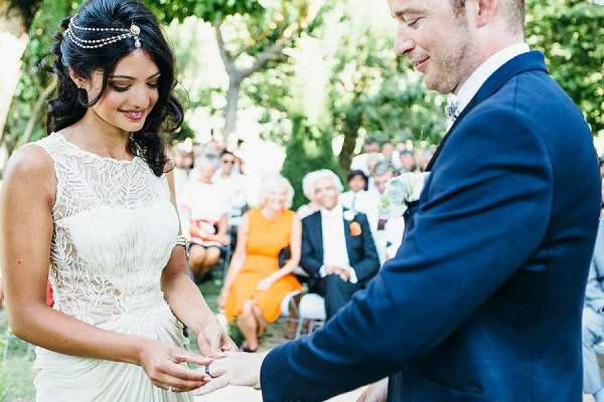 The couple is exchanging rings during an outdoor wedding ceremony, with the groom placing a ring on the bride's finger. The ceremony takes place in a shaded garden setting with seated guests visible in the background. The bride wears a cream/champagne embroidered sleeveless gown with a boho-inspired pearl and crystal draped headpiece worn across her forehead, while the groom wears a navy suit with a floral boutonniere. The shot is a medium close-up portrait focusing on the couple's hands and expressions, with guests softly out of focus behind them.