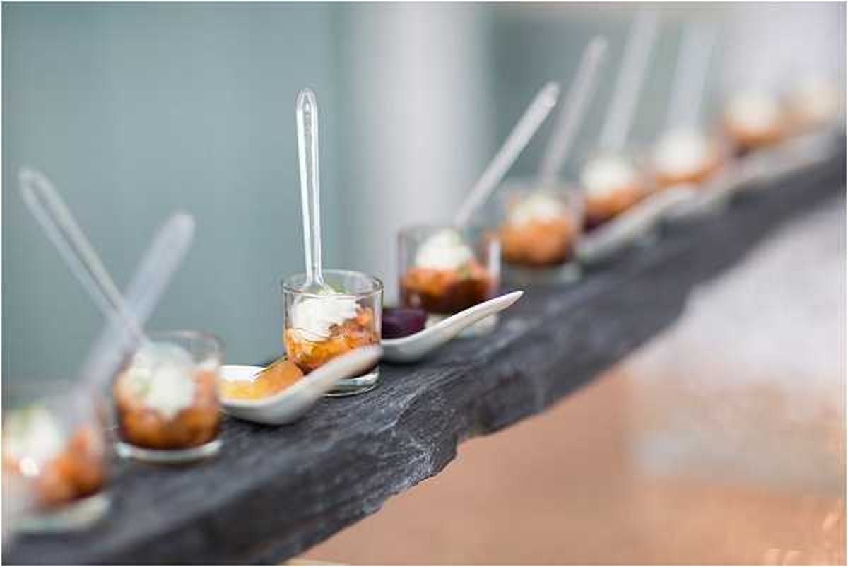 Close-up detail shot of wedding canapés or amuse-bouche arranged in a row along a dark slate serving board. The display includes white ceramic tasting spoons holding small bites topped with a cream or mousse garnish, alongside small glass shot cups containing layered ingredients with a white cream topping and clear cocktail picks. The food styling has a modern, refined catering aesthetic. The background is softly blurred in a muted teal-grey tone, keeping full focus on the food presentation in the foreground.