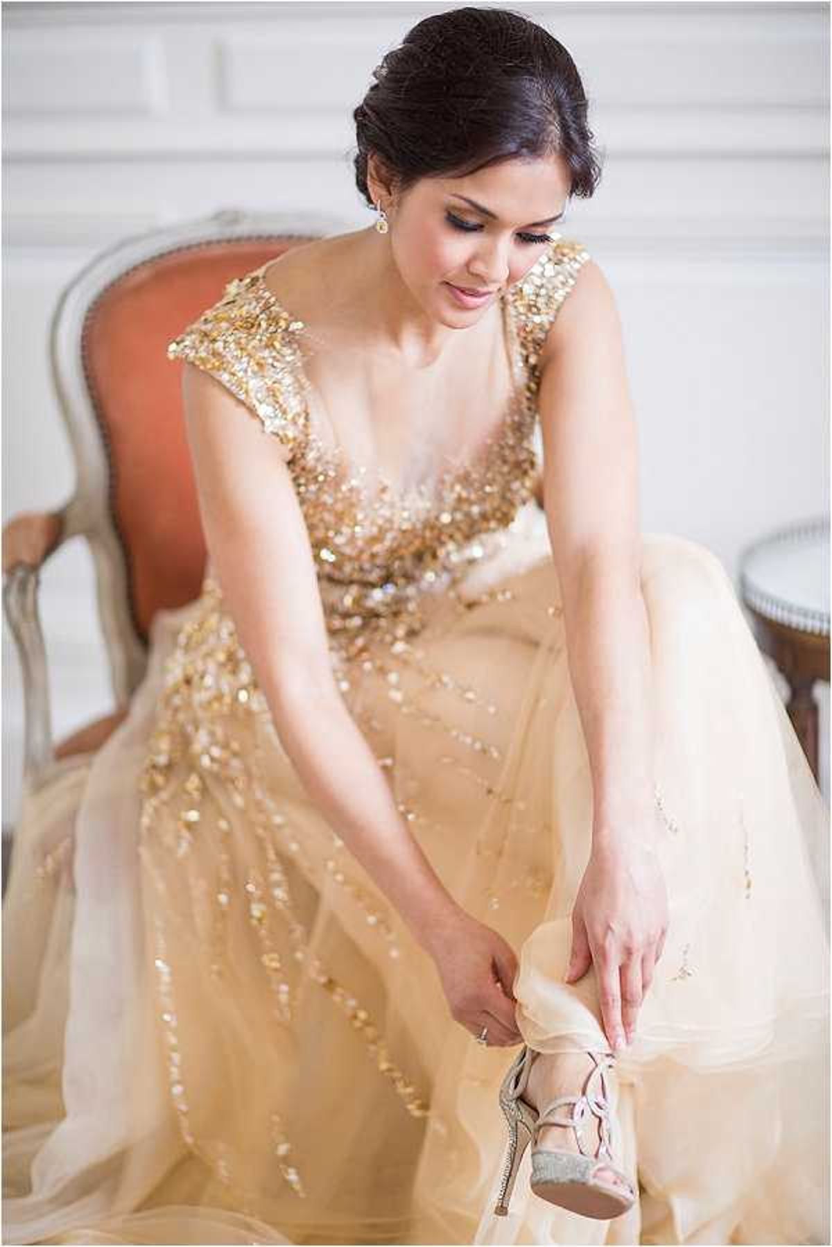 A bride is seated in a French-style armchair with a salmon-colored upholstered seat, fastening a strappy silver heeled sandal onto her foot. She is wearing a champagne and gold ball gown with a heavily sequined and beaded bodice and cap sleeves, and a layered tulle skirt with scattered gold embellishments. Her dark hair is styled in an updo, and she wears small drop earrings. The setting is an indoor room with white paneled walls, suggesting a classic interior. The composition is a mid-length portrait shot taken at a slight angle, with soft, even natural light creating a bright, airy feel.