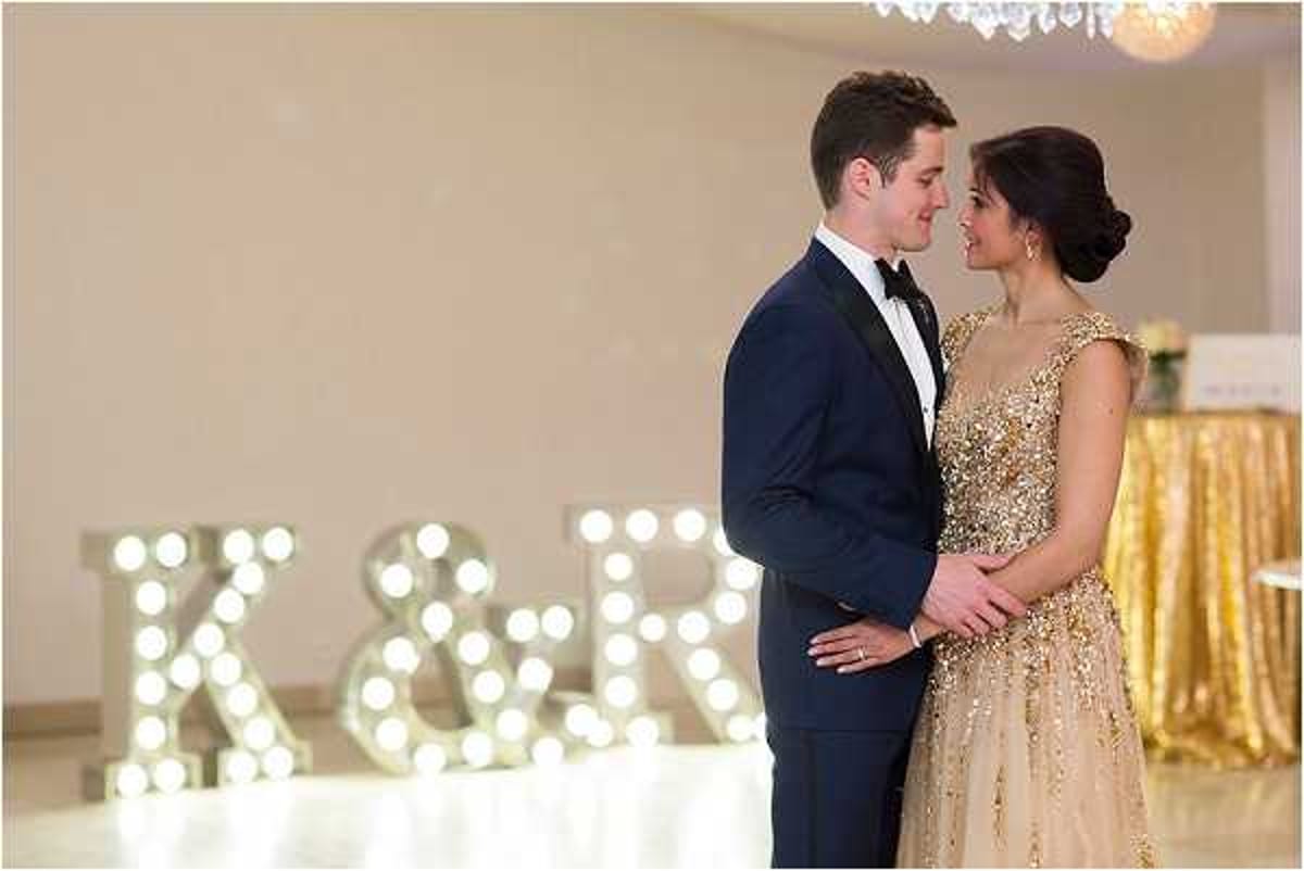 A couple poses together on a dance floor during what appears to be an indoor wedding reception in a ballroom or event hall. The groom wears a navy blue tuxedo with a black bow tie, while the bride wears a cap-sleeve gold sequined and beaded gown with a champagne base. Behind them are large illuminated marquee letters spelling 'K & R' with round bulb lighting, and to the right a table draped in a gold sequined tablecloth is partially visible, along with a crystal chandelier in the upper right corner. The decor palette is gold and navy, styled in a classic formal theme, captured in a mid-length portrait shot with the couple facing each other.