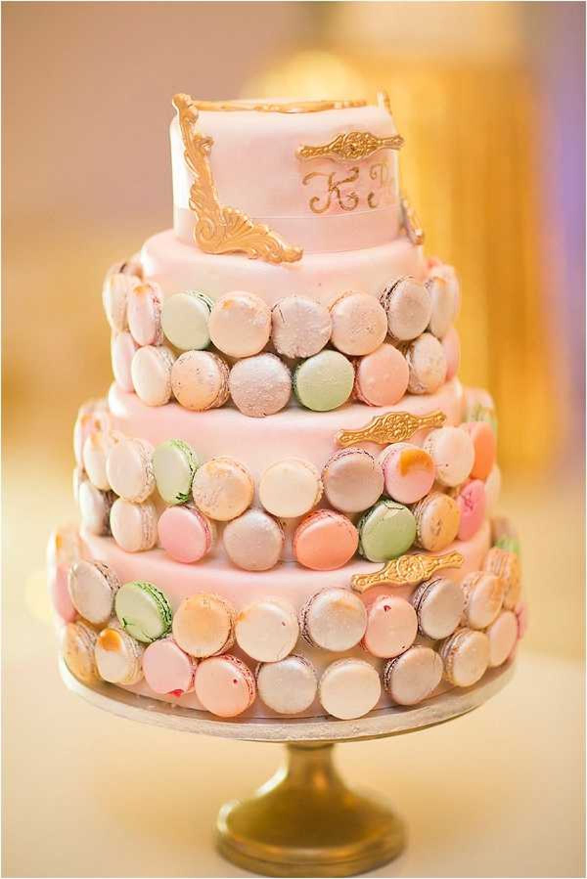 Close-up detail shot of a four-tiered wedding cake covered in pale pink fondant and decorated with rows of French macarons in pastel shades of pink, mint green, peach, and ivory. The top tier features gold ornate baroque-style corner decorations and a gold script monogram, while gold decorative accents are also placed between the macaron rows on the lower tiers. The cake is displayed on a gold pedestal cake stand. The background is softly blurred with warm golden and pale purple tones consistent with a reception venue interior.
