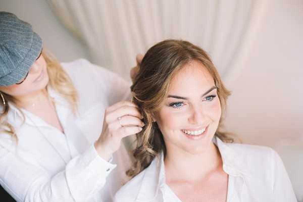 A getting-ready scene showing a bride seated while a hairstylist works on her loose, softly curled updo. The bride, wearing a white robe or shirt, has blue eyes and is smiling directly at the camera. The hairstylist, visible in the background, wears a grey flat cap and a white shirt. The setting appears to be a bright, softly lit indoor room with neutral-toned curtains. The shot is a close-up portrait with a shallow depth of field, keeping focus on the bride's face and hair.