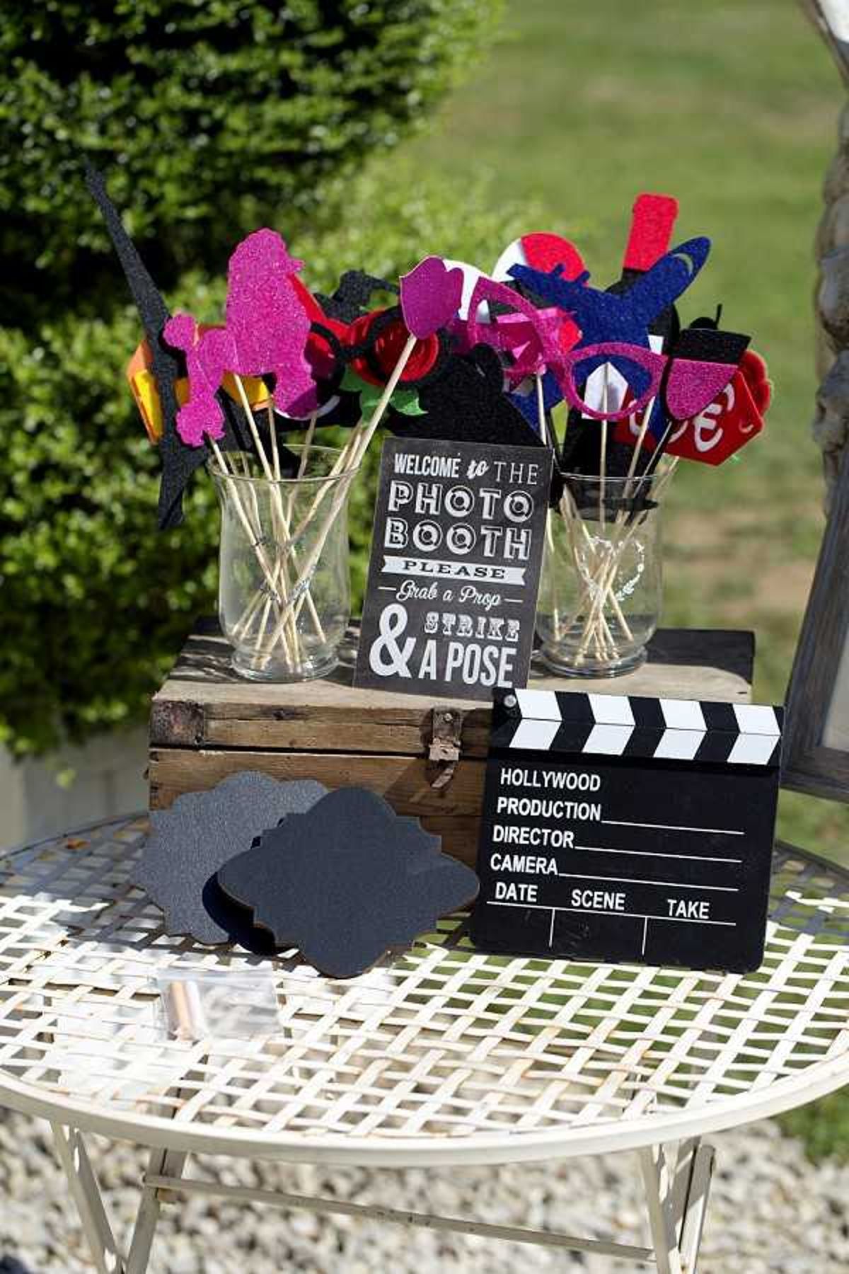 A photo booth prop station set up outdoors on a white wrought-iron garden table, photographed as a detail close-up shot. Two clear glass jars hold an assortment of glittery stick props in hot pink, black, red, navy blue, and gold, including shapes such as lips, hats, glasses, and a Paris Eiffel Tower silhouette. A chalkboard-style sign reads 'Welcome to the Photo Booth – Please Grab a Prop & Strike a Pose,' and a Hollywood-style film clapperboard prop sits at the front of the table alongside dark grey decorative chalkboard speech-bubble cutouts. The props are displayed on top of a rustic wooden storage box, suggesting a Hollywood or cinema-themed wedding reception decor concept. The setting appears to be an outdoor cocktail hour or reception area.