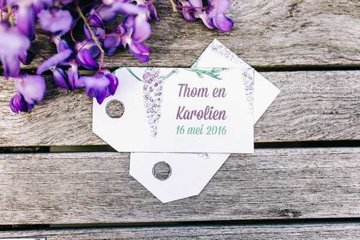Close-up flat lay detail shot of two white wedding favor tags printed with 'Thom en Karolien 16 mei 2016' in purple and teal typography, decorated with a watercolor wisteria illustration in shades of purple and green. The tags are fastened with small silver brad fasteners and arranged overlapping on a weathered grey wooden surface. Fresh purple wisteria clusters are placed in the upper left corner, echoing the printed floral motif on the tags. The overall decor palette centers on purple and teal with a botanical theme.