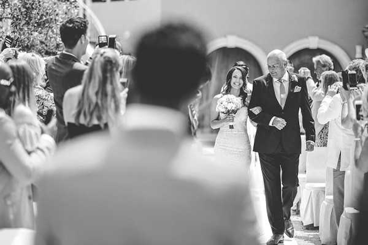 A black-and-white image of a bride walking down the aisle accompanied by an older man, likely her father, during an outdoor ceremony. The bride is wearing a lace gown and carrying a round bouquet, and she is smiling as she approaches. The ceremony is set in what appears to be a courtyard or terrace with arched architectural features visible in the background, with white chairs lining the aisle and approximately 20–30 seated and standing guests visible on both sides, several of whom are photographing the procession. The shot is taken from behind the groom in the foreground, creating a point-of-view perspective with shallow depth of field that places the approaching bride in sharp focus against the softly blurred crowd.