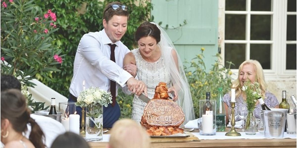 The bride and groom are cutting into a traditional French croquembouche or profiterole tower wedding cake at an outdoor reception, with the cake decorated with a wooden sign reading 'pain' and 'mirage' (or similar lettering). The bride wears a white lace or embroidered gown with a veil, while the groom wears a white dress shirt with a burgundy tie and dark trousers. The reception table is styled with white pillar candles in gold candlesticks, small glass bud vases with greenery and small white blooms, wine bottles, and a champagne ice bucket, set on a wooden farm table. A female guest is visible laughing in the background, and the setting is an outdoor garden terrace with lush green foliage behind. This is a medium-wide shot capturing the cake-cutting moment with guests seated in the foreground.