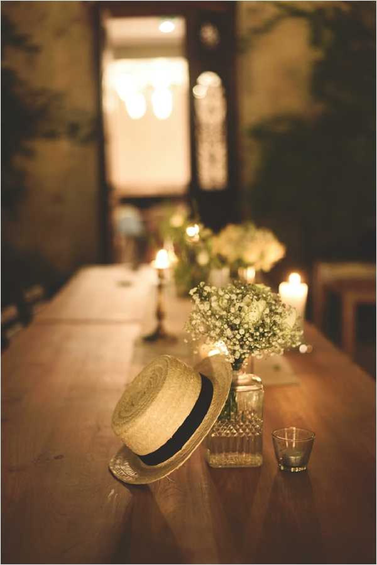 Close-up detail shot of a rustic wooden reception table decorated in a vintage style. In the foreground, a straw boater hat with a navy ribbon band rests against a cut-crystal vase holding baby's breath, alongside a small glass votive candle holder. Further along the long table, additional arrangements of white flowers and baby's breath are flanked by pillar candles and brass candlesticks, creating a warm, candlelit atmosphere. The setting appears to be an indoor courtyard or stone-walled room, with a lit doorway visible in the blurred background, reinforcing a rustic, vintage-inspired decor palette of natural textures, warm amber candlelight, and white blooms.