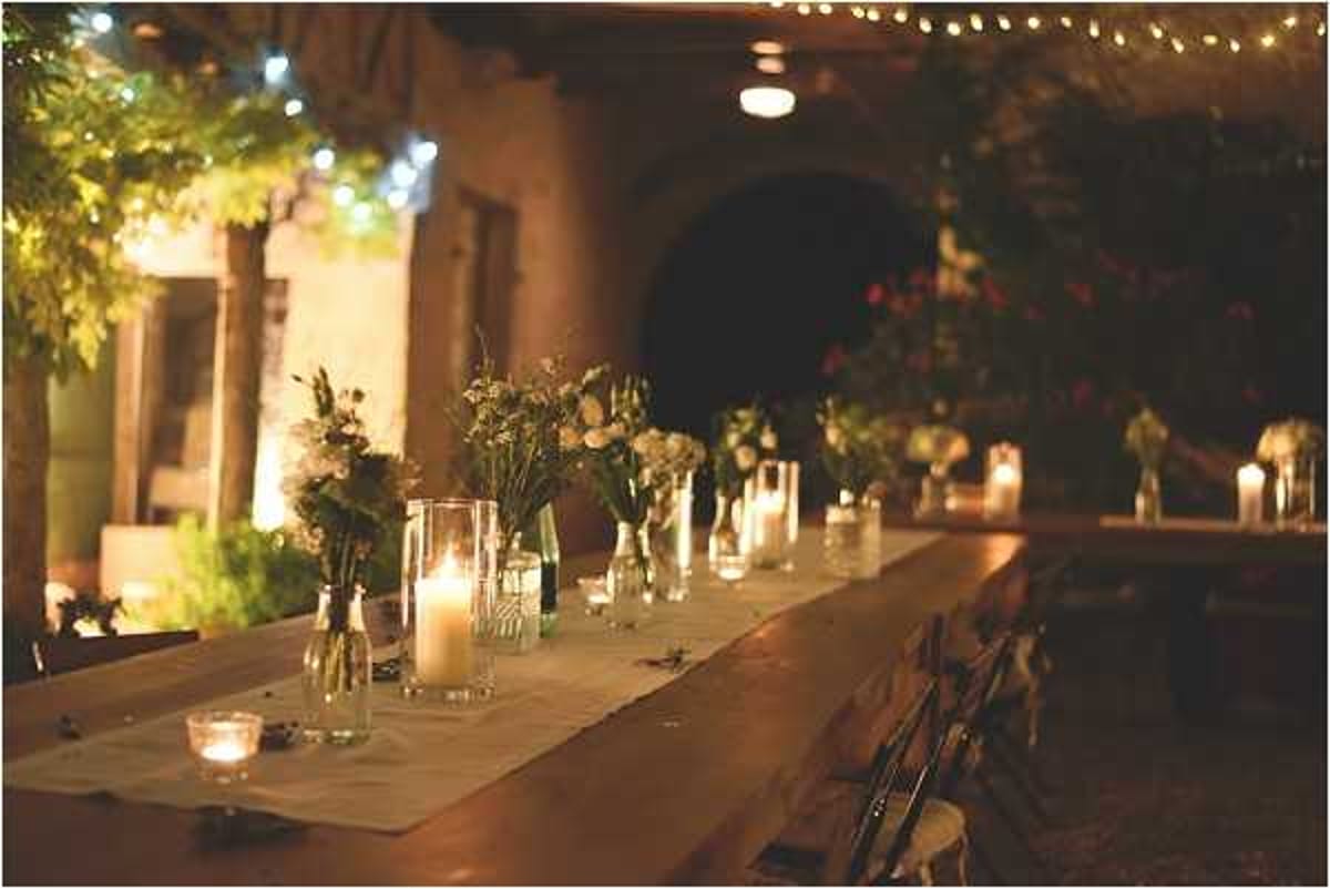 An evening outdoor reception dinner table set up in what appears to be a rustic courtyard or covered terrace, with stone archways visible in the background. The long wooden farm-style table is styled with a linen runner, pillar candles and votive candles in glass holders, and small bud vases containing loosely arranged white and green wildflower-style blooms. Overhead string bistro lights add warm ambient lighting across the space, and folding chairs are visible along the sides of the table. The overall decor palette is warm and rustic, with an intimate, candlelit atmosphere.