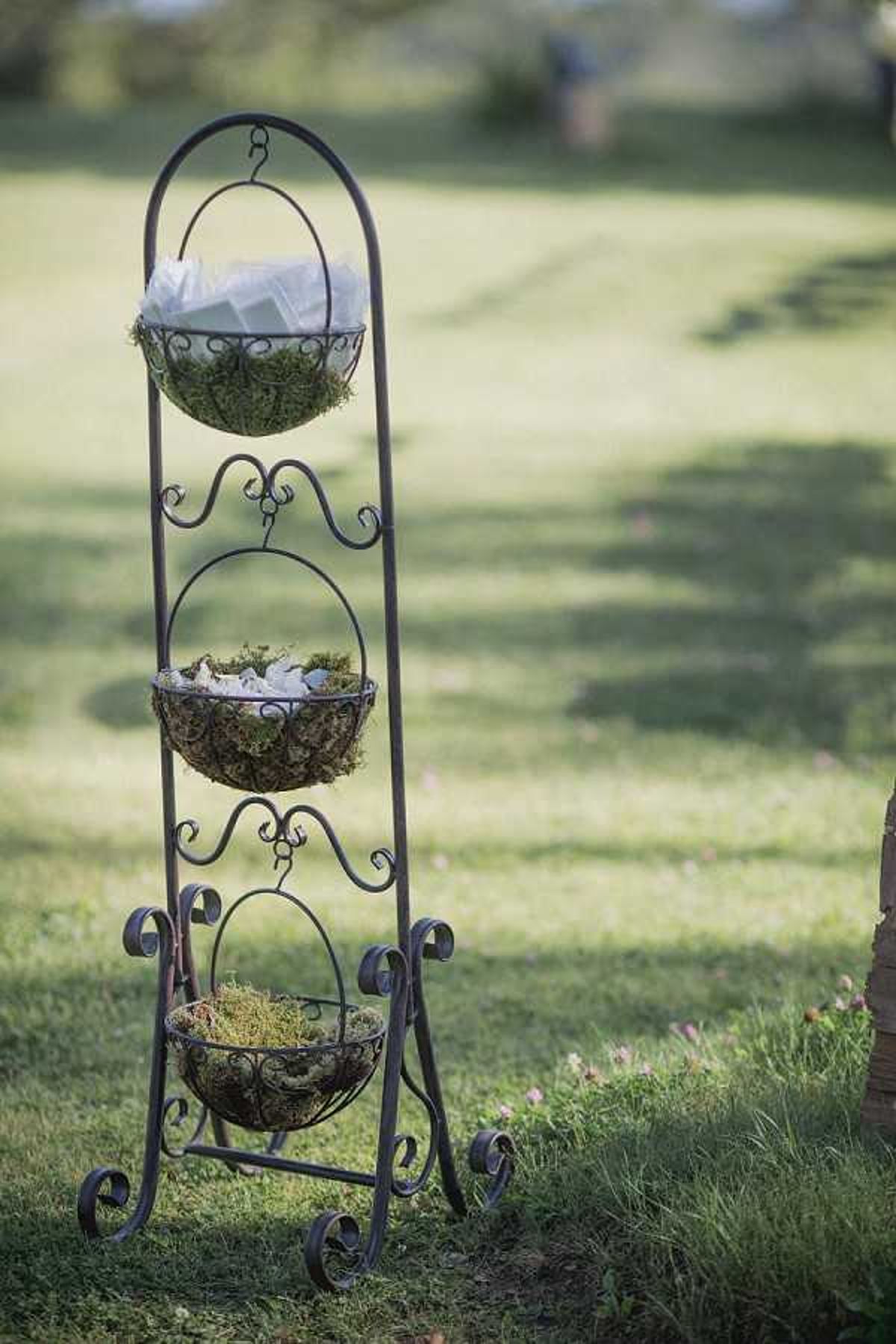 A close-up detail shot of a decorative wrought iron three-tiered hanging basket stand placed on a lawn at an outdoor wedding. The ornate dark metal stand features scrollwork detailing and holds three wire baskets lined with green moss; the upper baskets contain what appear to be small white paper or fabric favor packets for guests, while the bottom basket holds moss only. The styling is rustic-garden in theme, and the stand is positioned at the edge of what appears to be a ceremony or reception area on a grass lawn with soft bokeh background.