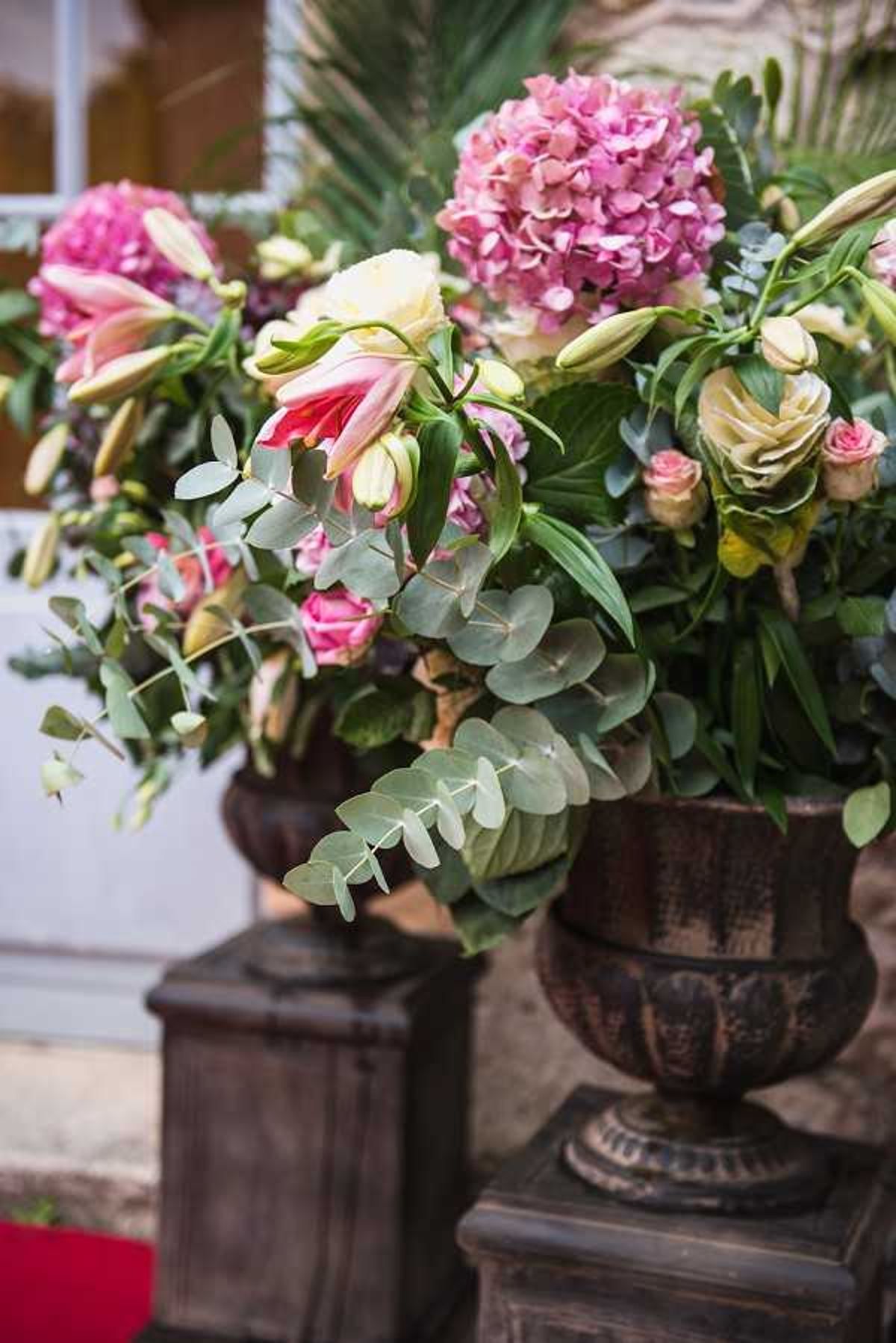 Close-up detail shot of two large floral arrangements displayed in dark bronze-toned ornate pedestal urns, positioned outdoors at what appears to be a venue entrance. Each urn contains a full, abundant arrangement featuring pink hydrangeas, blush and pink oriental lilies, cream and pink spray roses, cream lisianthus buds, and abundant eucalyptus foliage. The color palette is warm pink and blush tones with lush greenery. A red carpet is partially visible at the base left, and a building facade is softly blurred in the background.